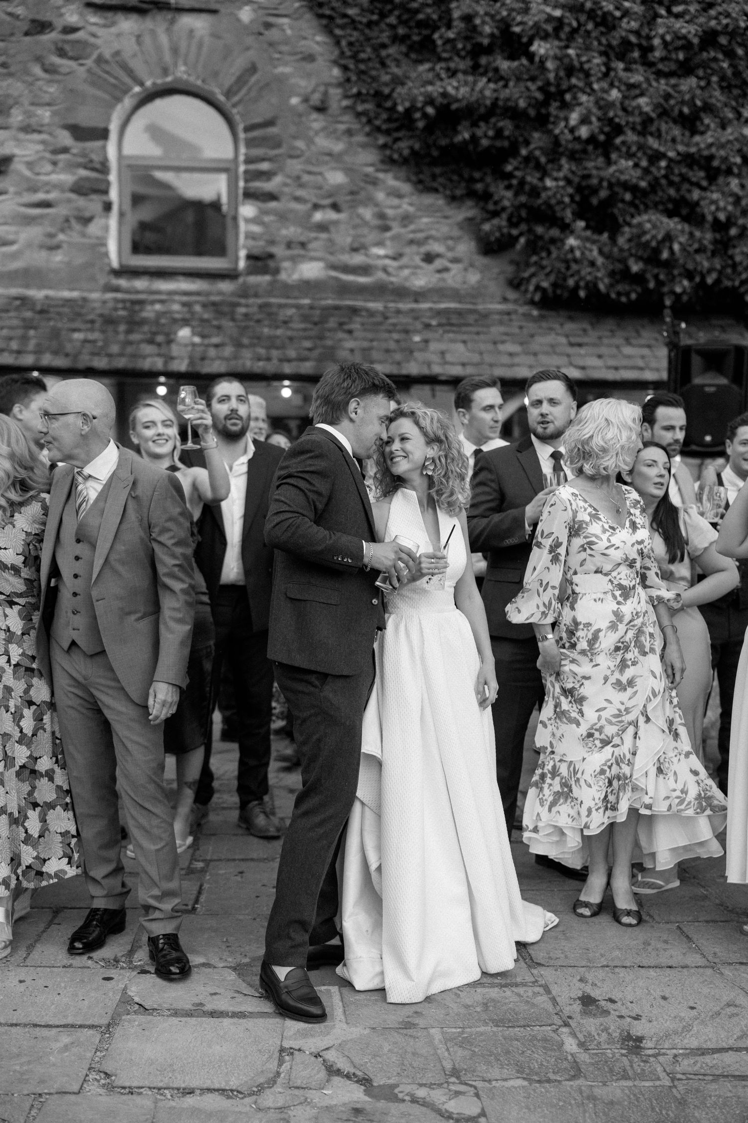 Black and white photo of wedding guests celebrating on a stone patio near a historic building.