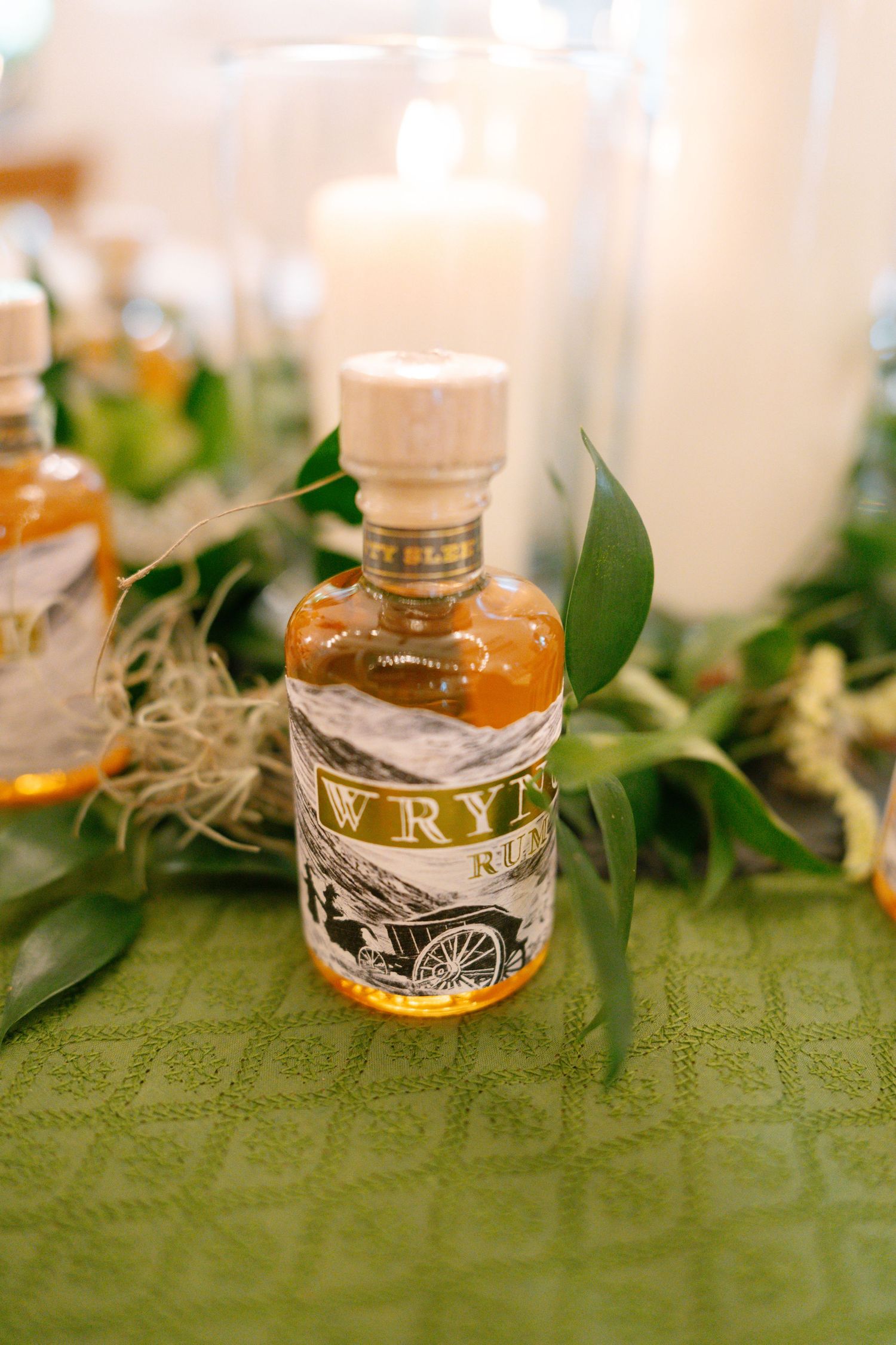 Decorative glass bottles with greenery on green tablecloth at wedding reception.
