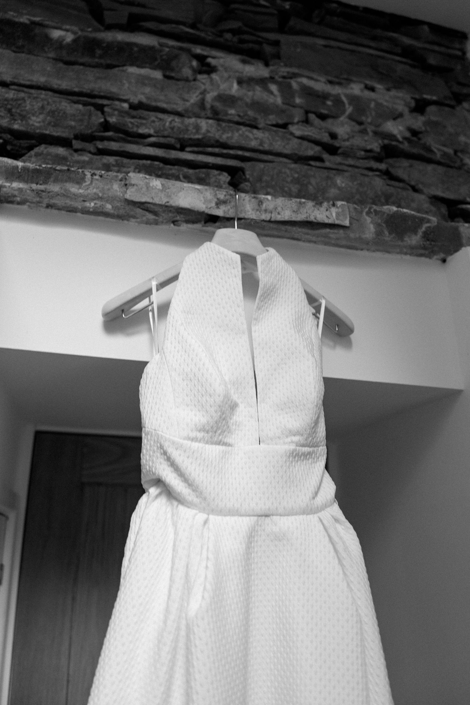 A white polka dot wedding dress hangs from a rustic wooden beam against a stone wall.