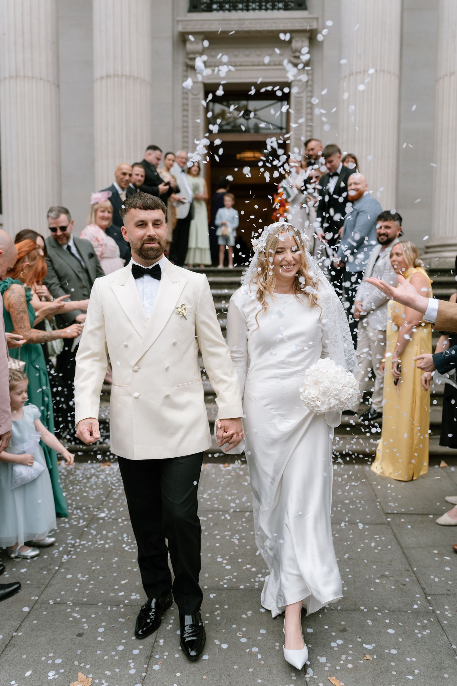 Newlyweds exit their wedding ceremony through a shower of confetti while wearing elegant white attire.