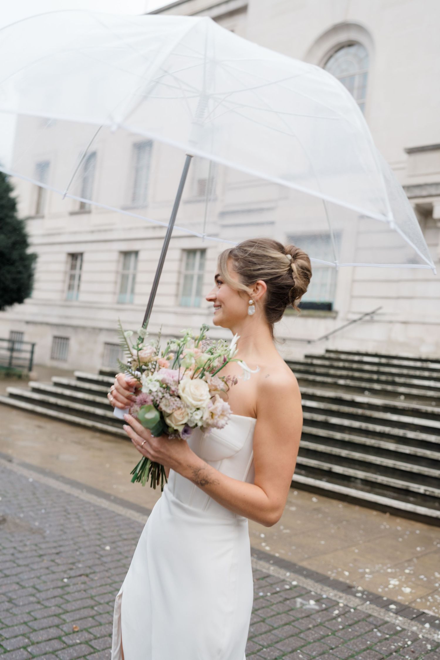 A bride in a strapless white wedding dress holds a clear umbrella and bouquet while standing on stone steps in the rain.