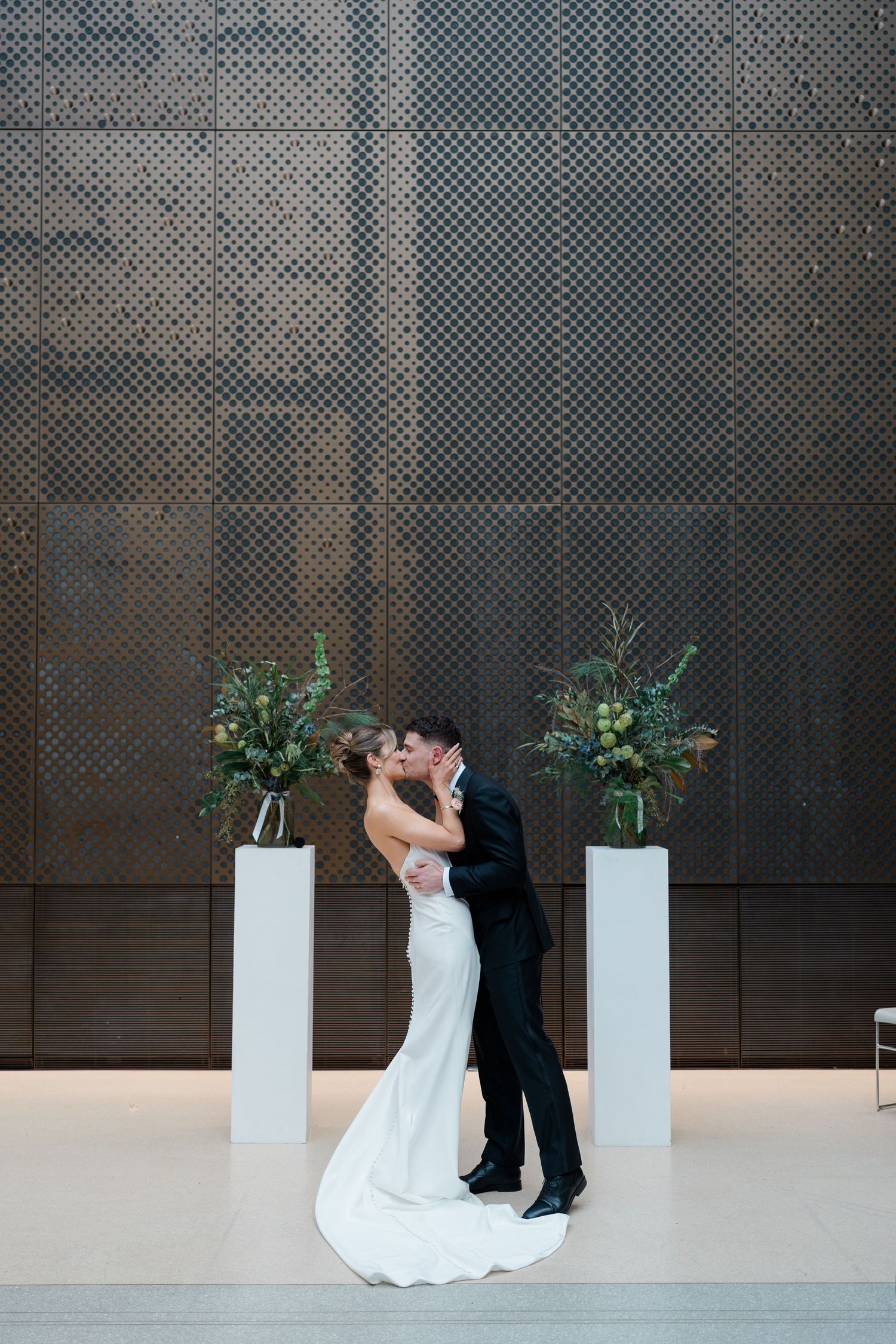A romantic dip pose sequence between a couple in wedding attire against an industrial concrete wall with white planters.