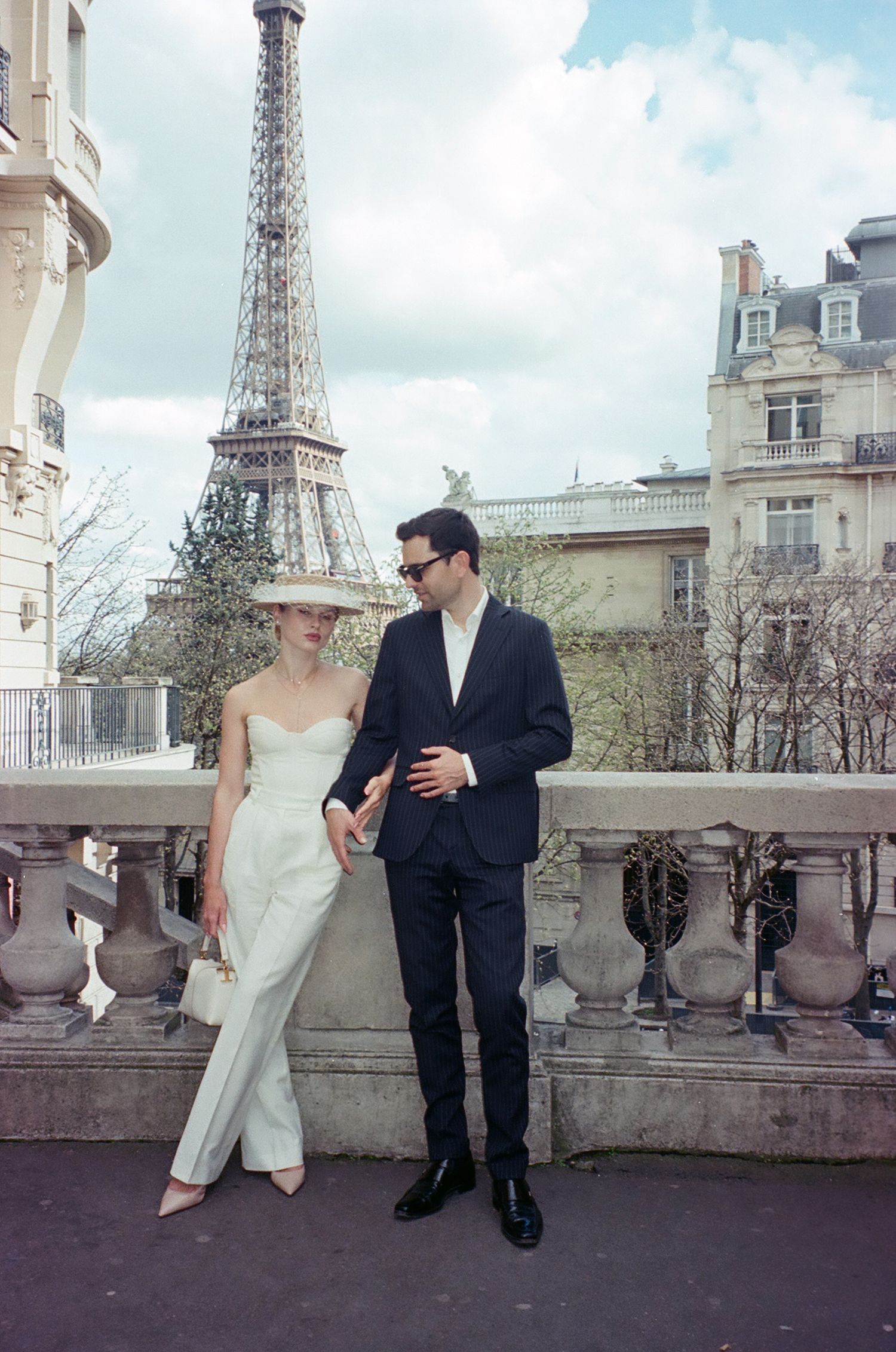 A romantic couple in formal attire poses together on a balcony with the iconic Eiffel Tower visible in Paris, France.