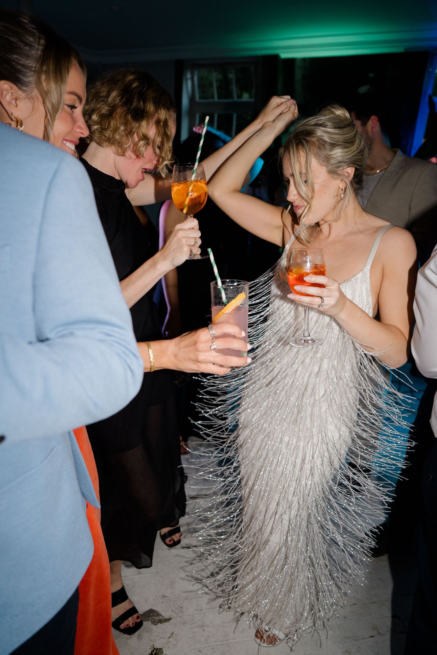 A bride in a fringed dress dances and celebrates at a reception with colorful lighting.