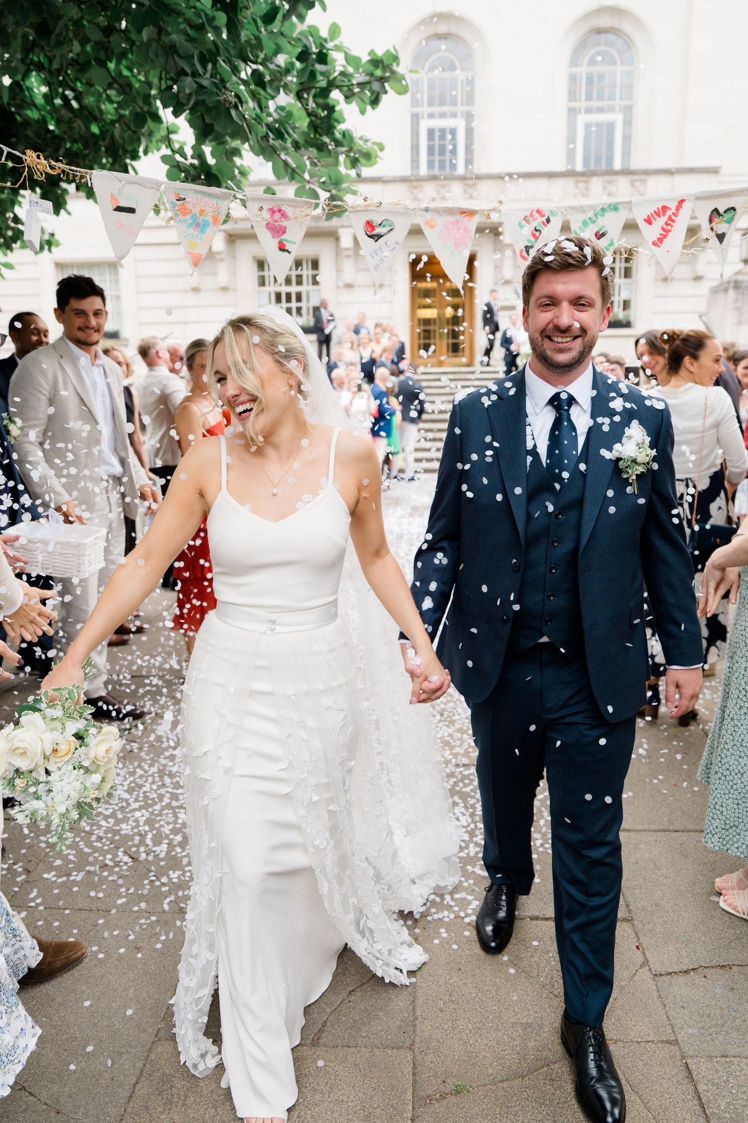A joyful wedding couple exits their ceremony while guests shower them with confetti on a grand building's steps.