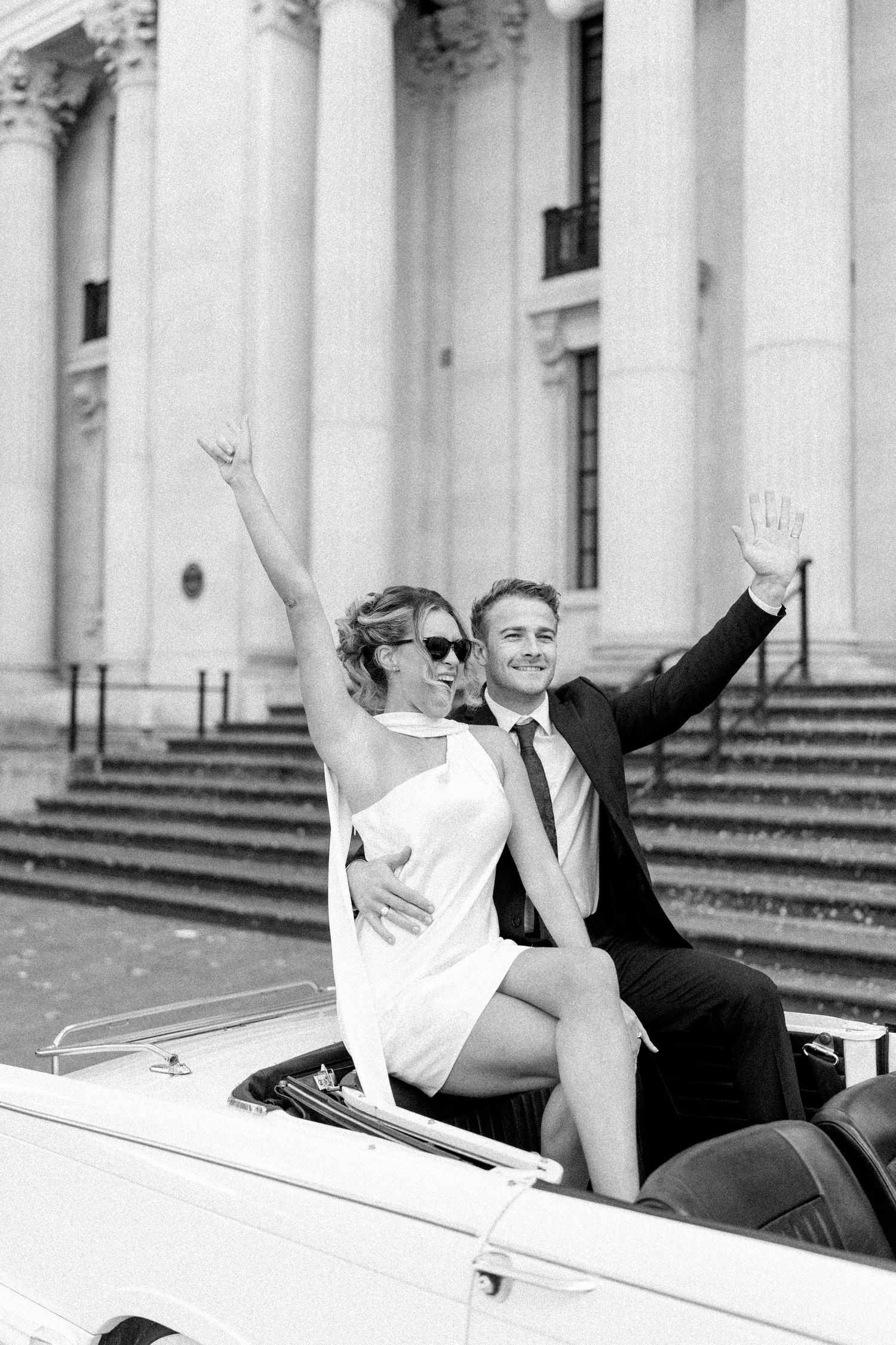 Joyful couple celebrates in classic white convertible car in front of historic building.