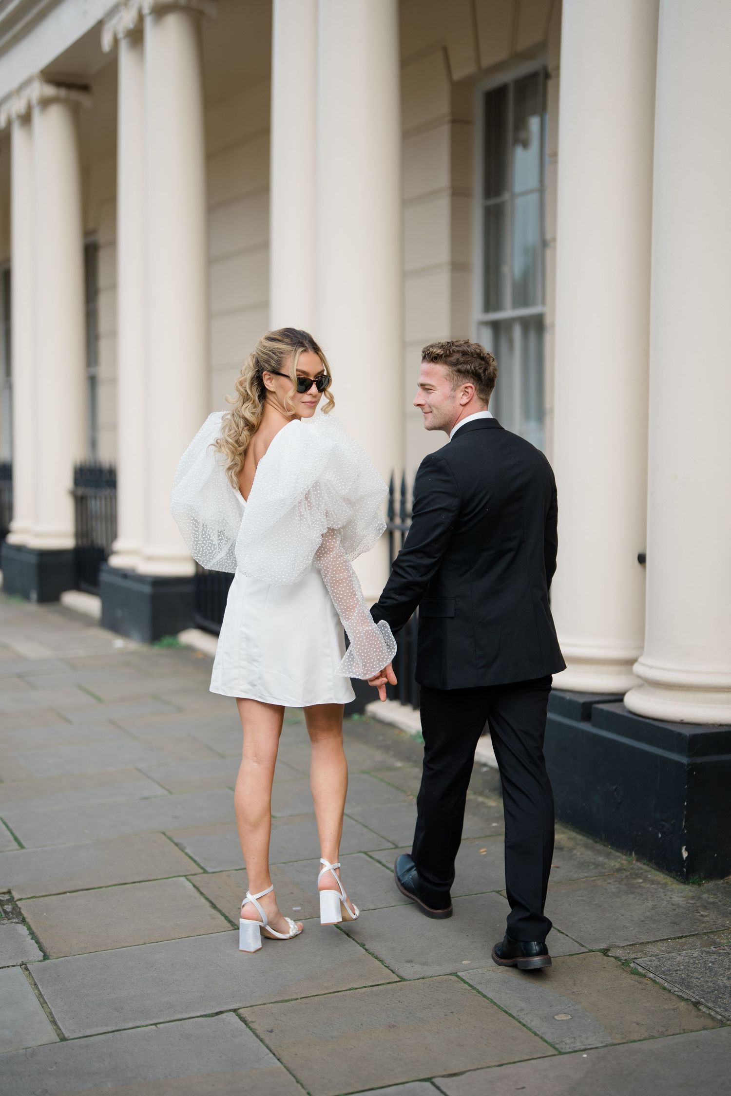 A couple in formal attire poses playfully on a stone walkway near classical columns.