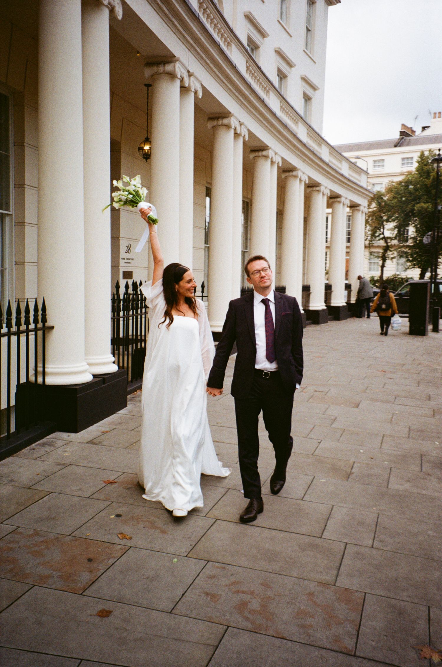 A wedding couple walks playfully along an elegant colonnade with white pillars and stone paving.