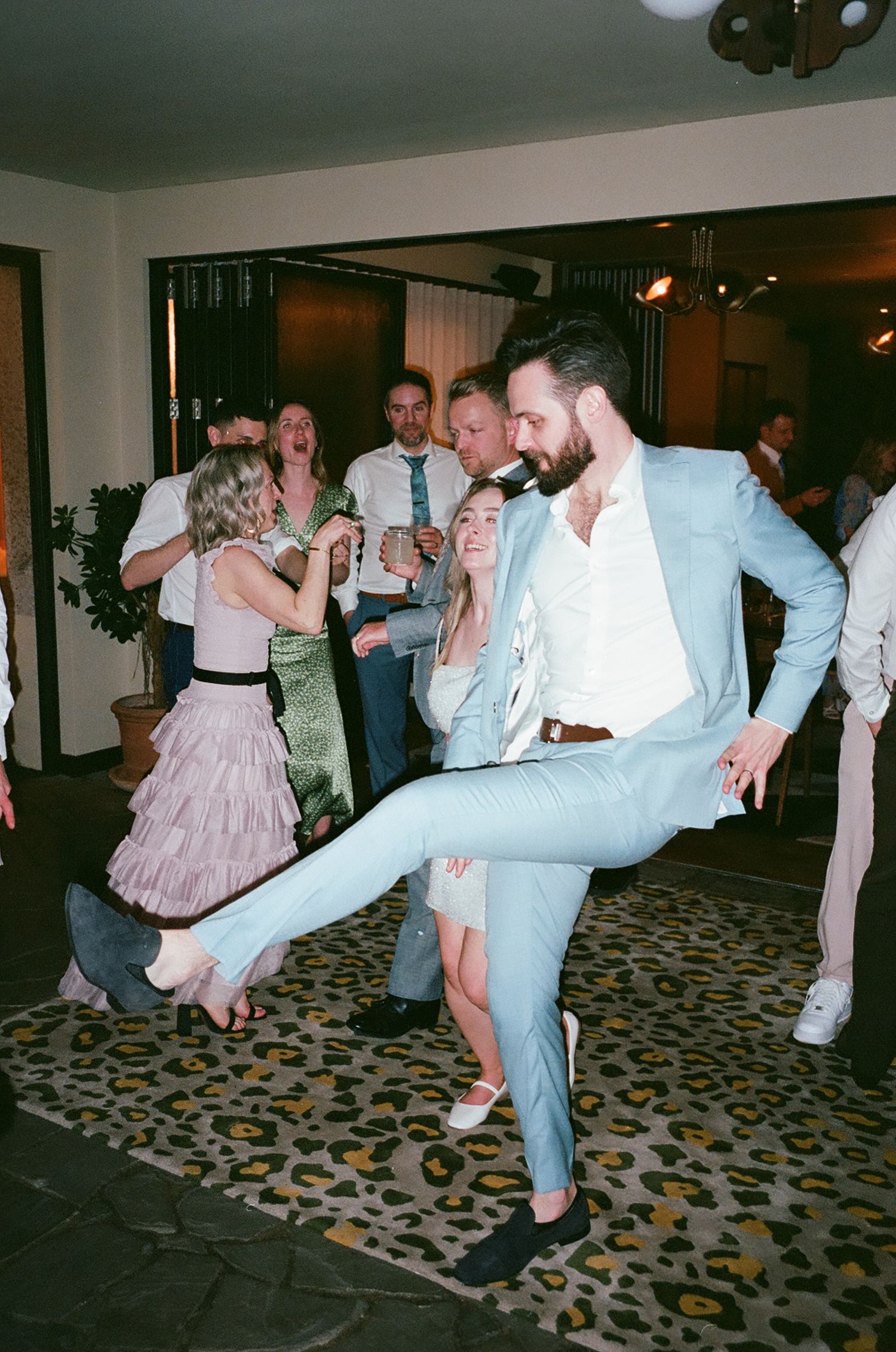 Party guests dancing and celebrating on a patterned carpet at an indoor event with retro film camera aesthetic.