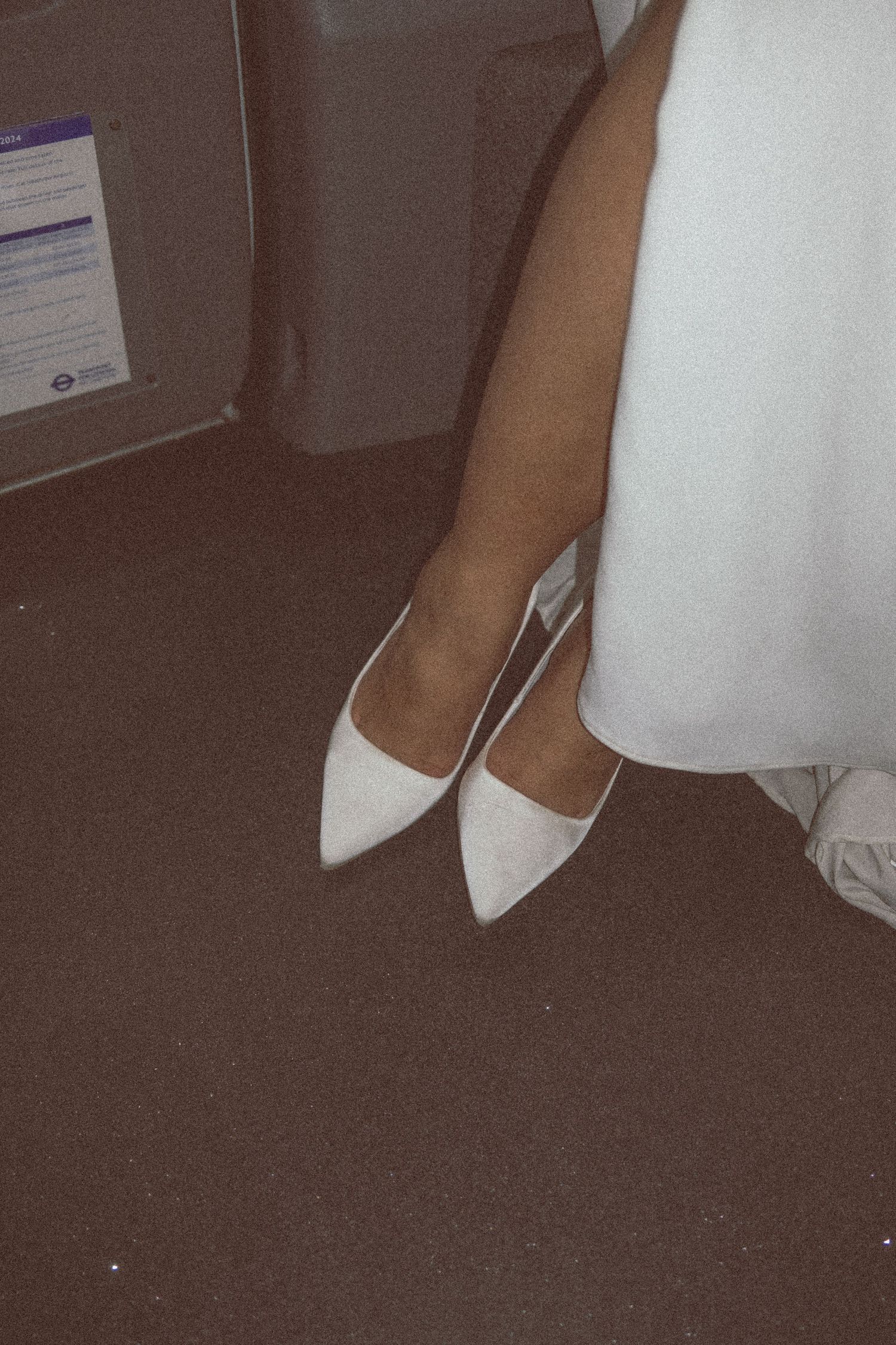 A pair of elegant white pointed-toe flats worn with a flowing white dress against a dark carpet.