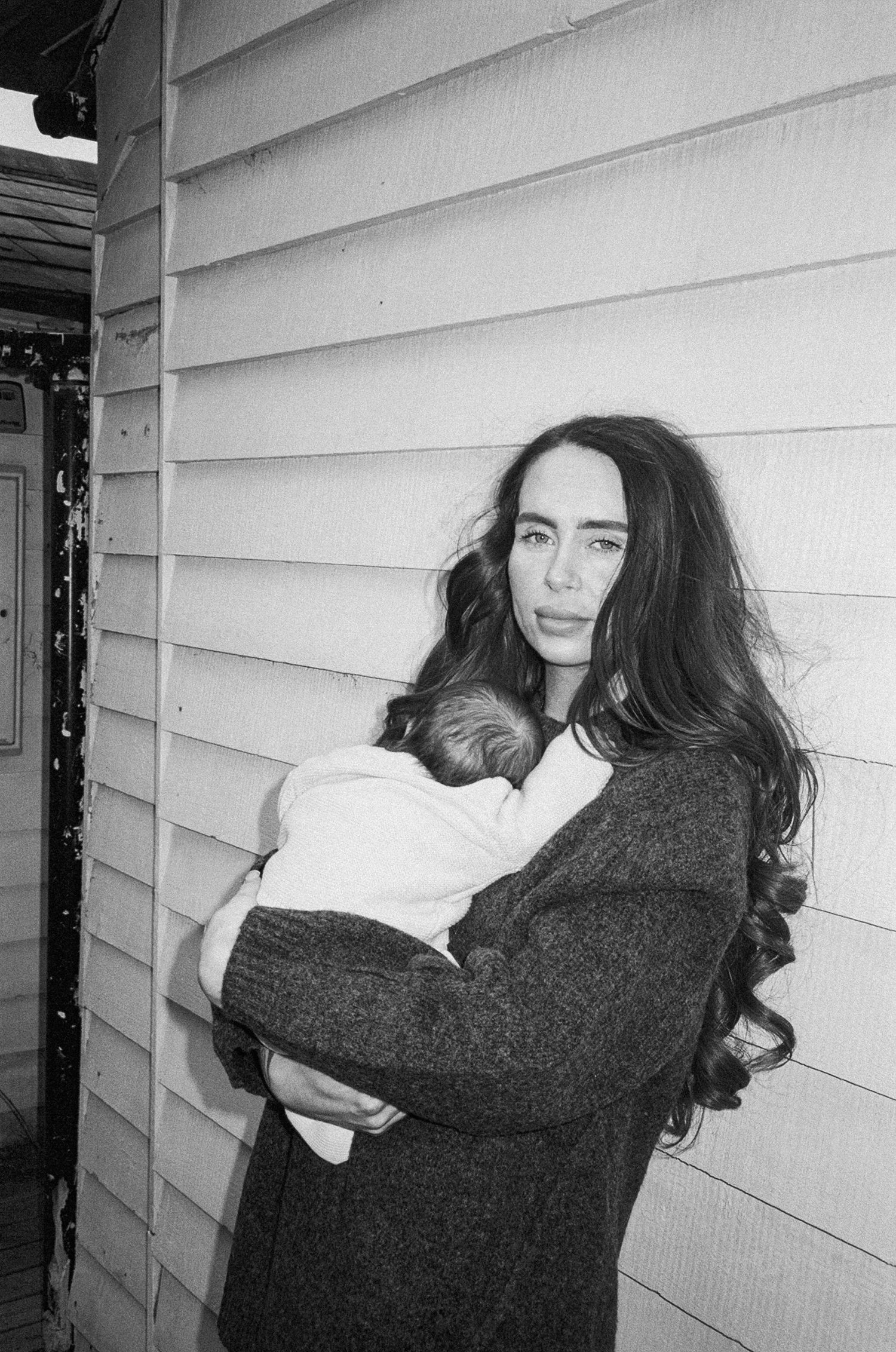 Black and white photo of a young woman with long dark hair holding a newborn baby against a wooden siding wall.