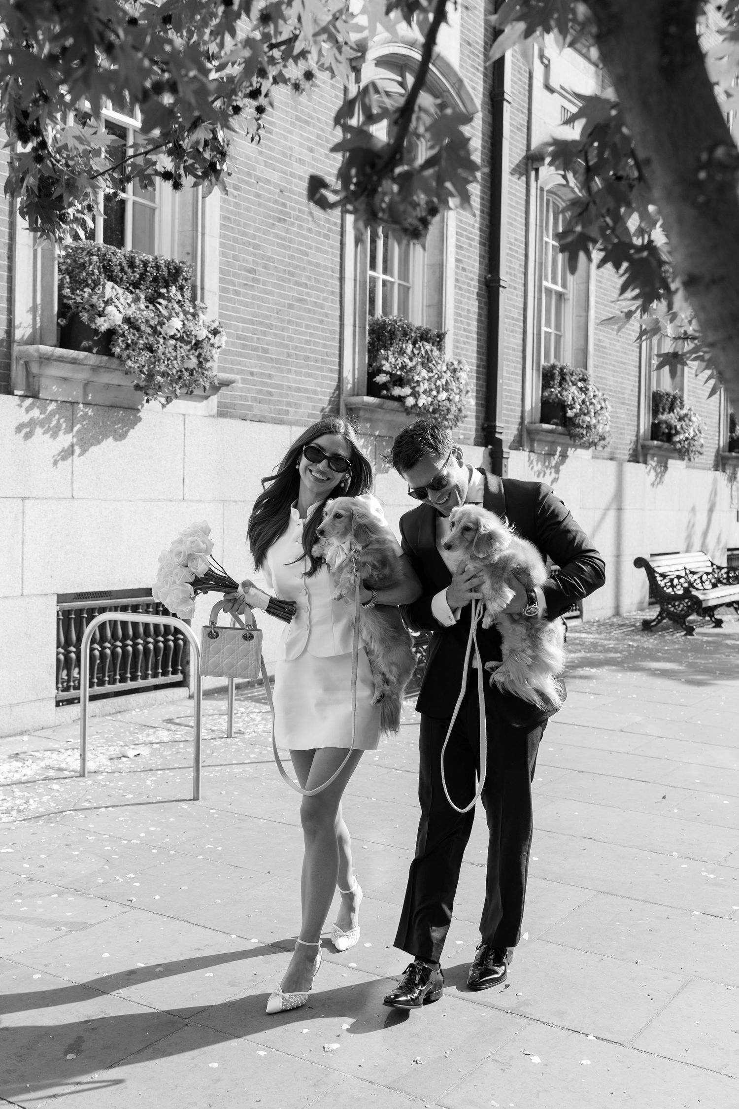 Black and white photo of newlyweds each holding a dachshund puppy on sunny city street.