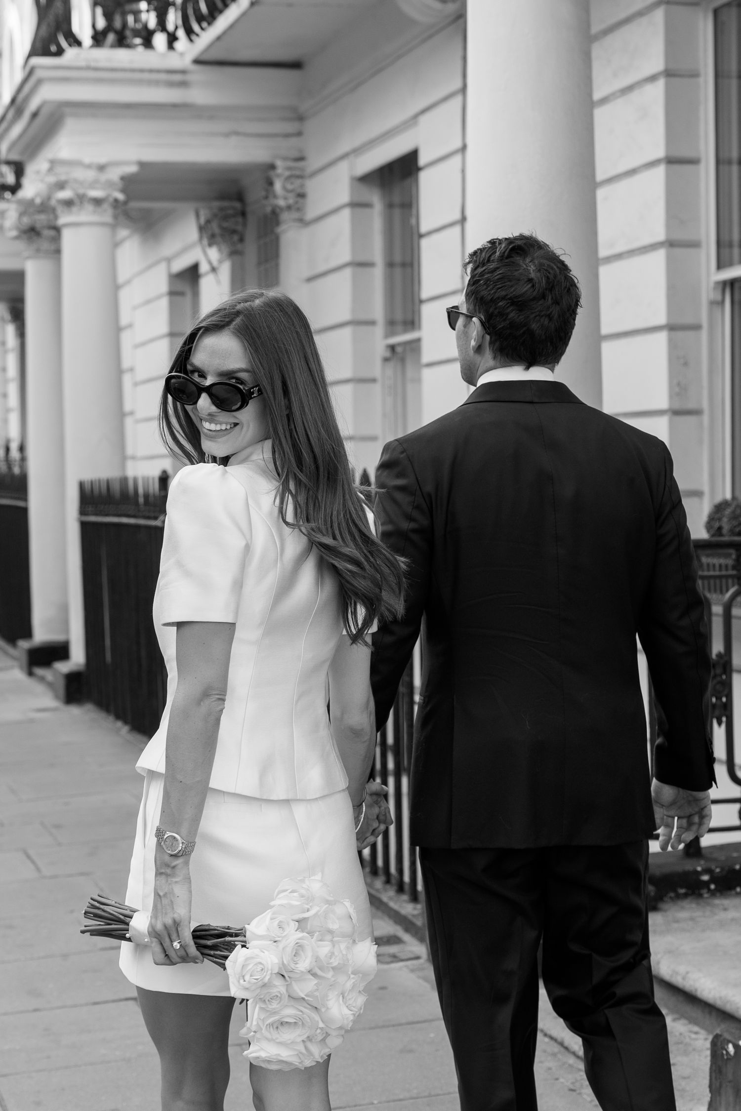 Smiling bride in white mini dress glances back while walking with groom.