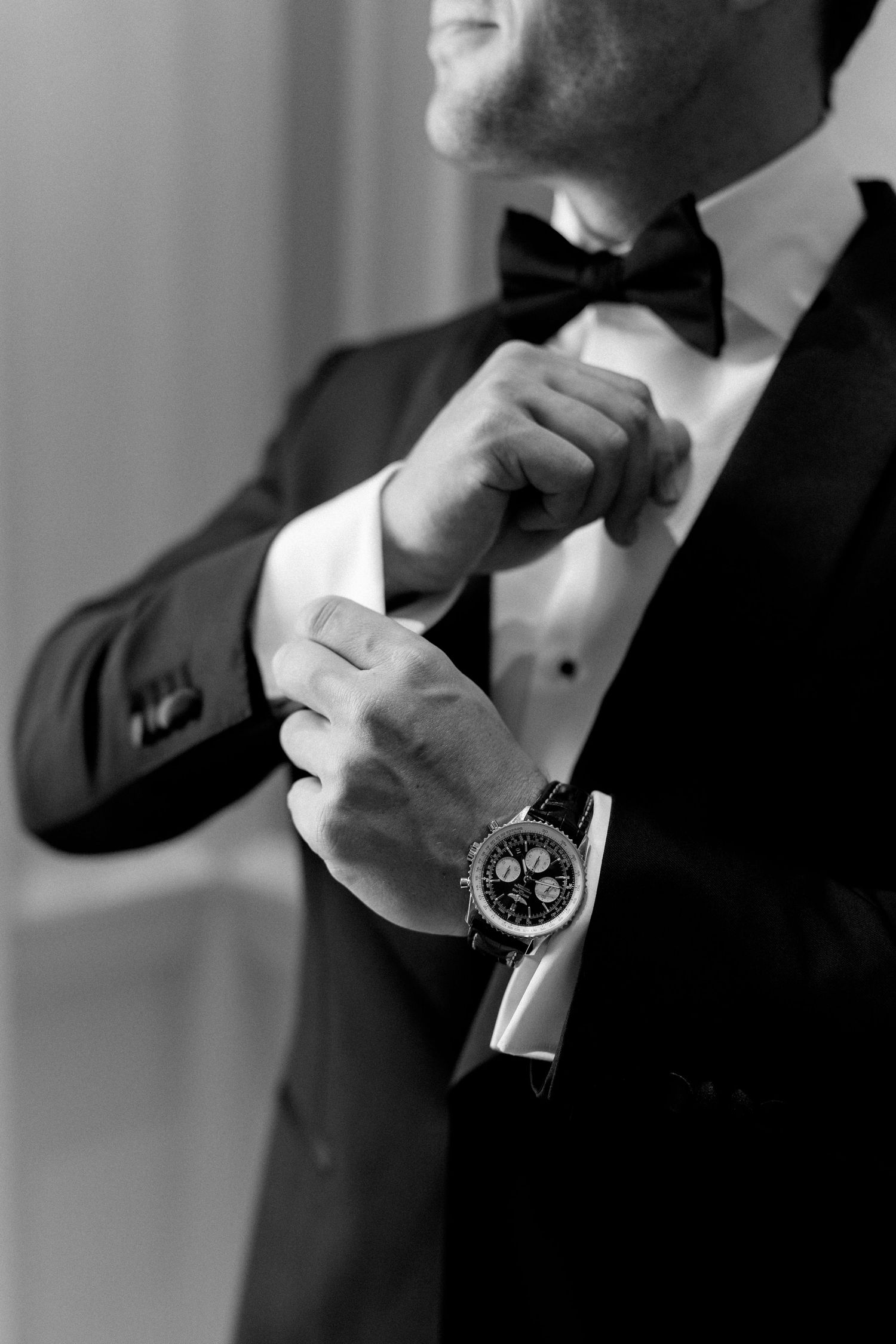 Groom in black tuxedo adjusts bow tie while wearing luxury chronograph watch, black and white photo.