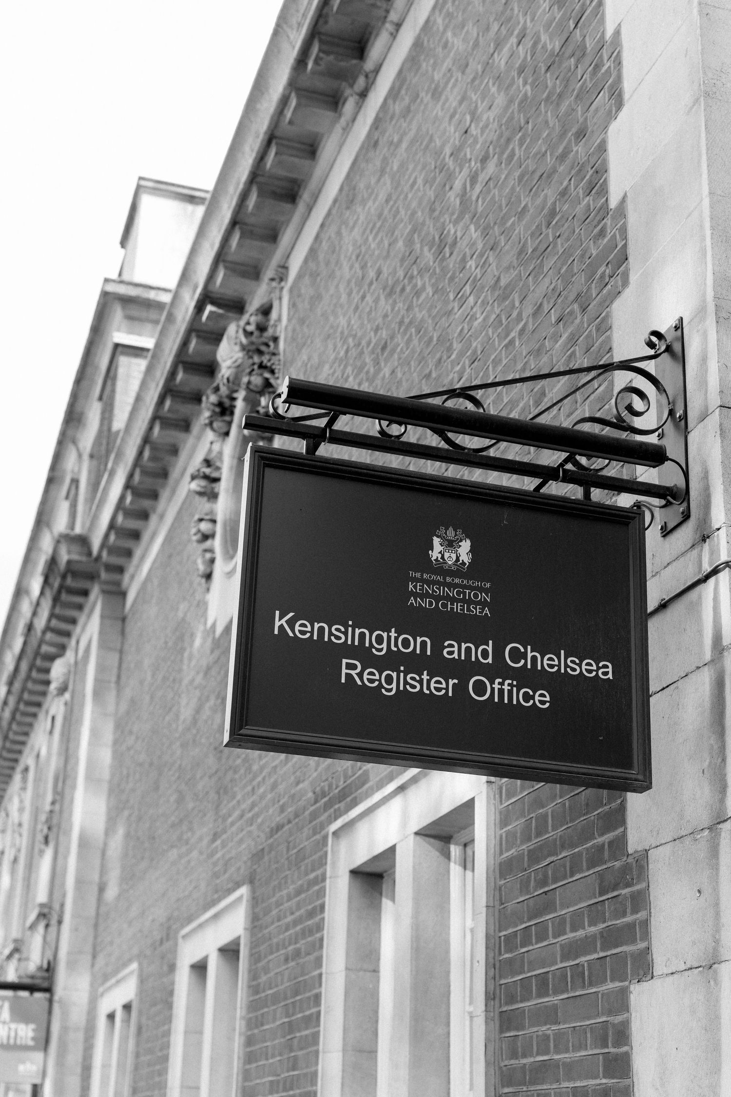 Black and white sign for Kensington and Chelsea Register Office mounted on brick building wall.
