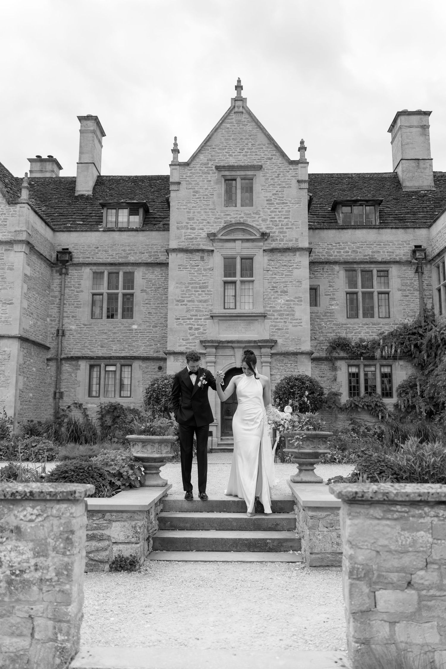 Bride and groom hold hands ascending stone steps before grand English manor in black and white.