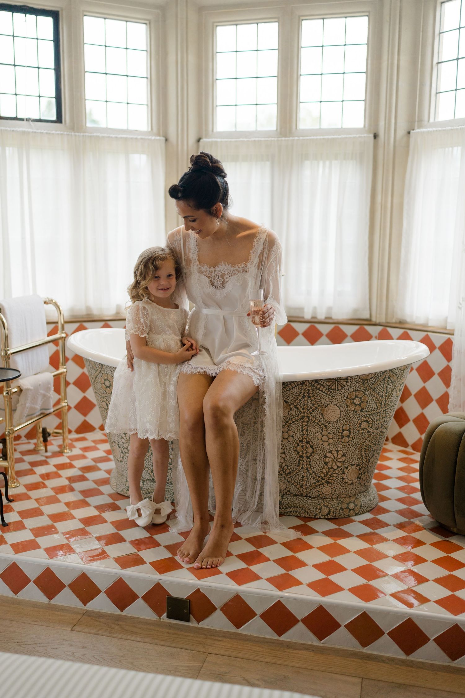 Bride in lace robe sitting on ornate bathtub edge with flower girl on orange diamond-tiled bathroom floor.