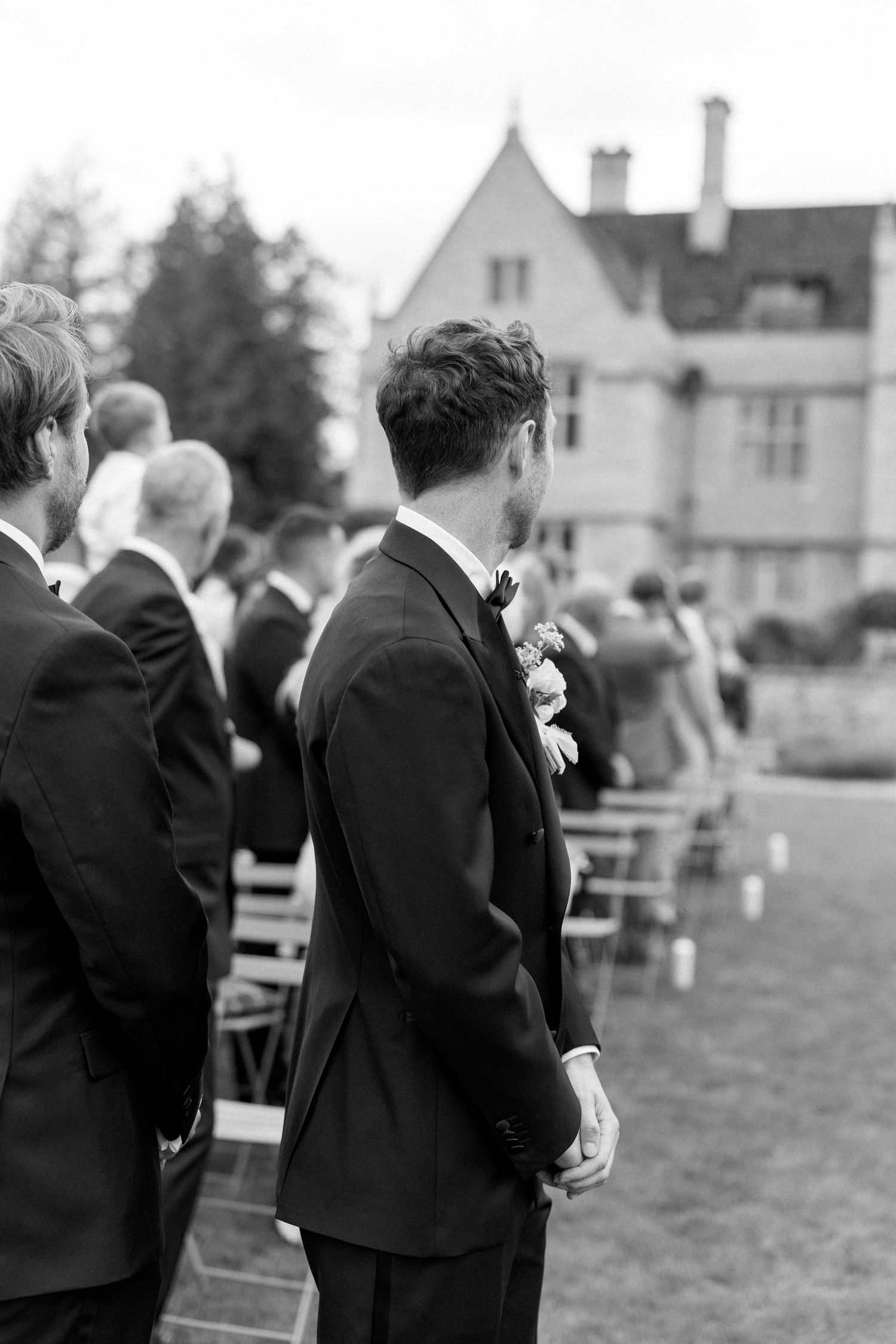 Black and white photo of groom in tuxedo awaiting bride at outdoor wedding ceremony.