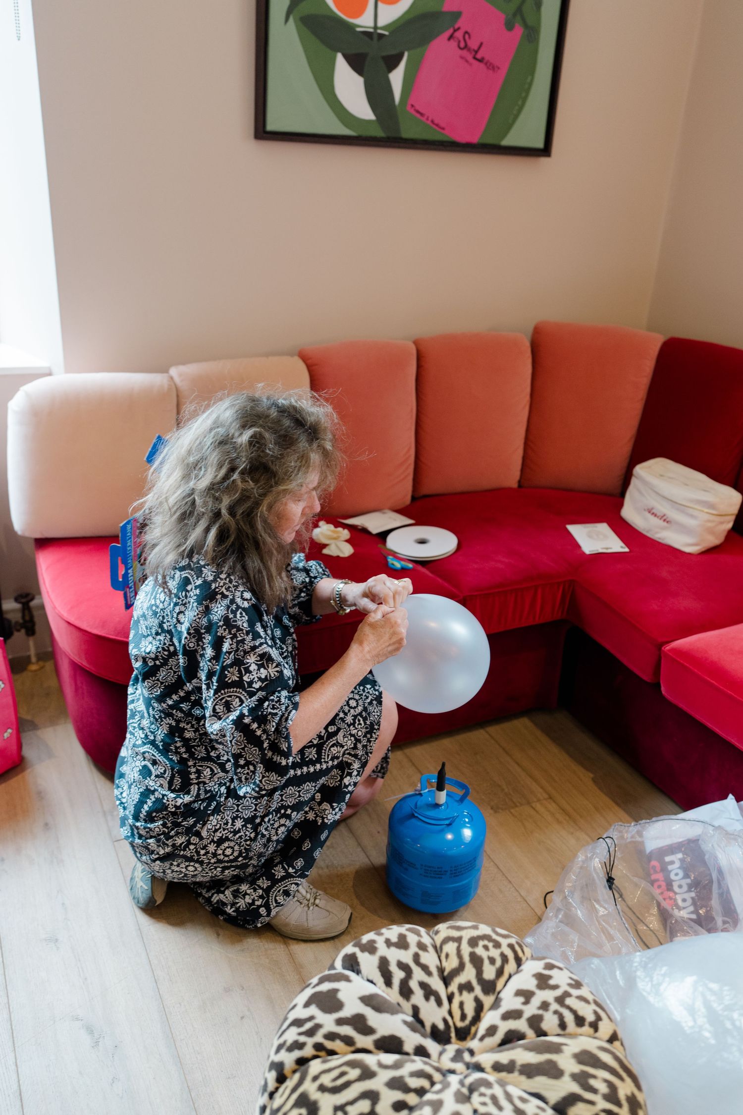 Woman kneeling on floor inflating balloon with helium tank near red sofa in living room.