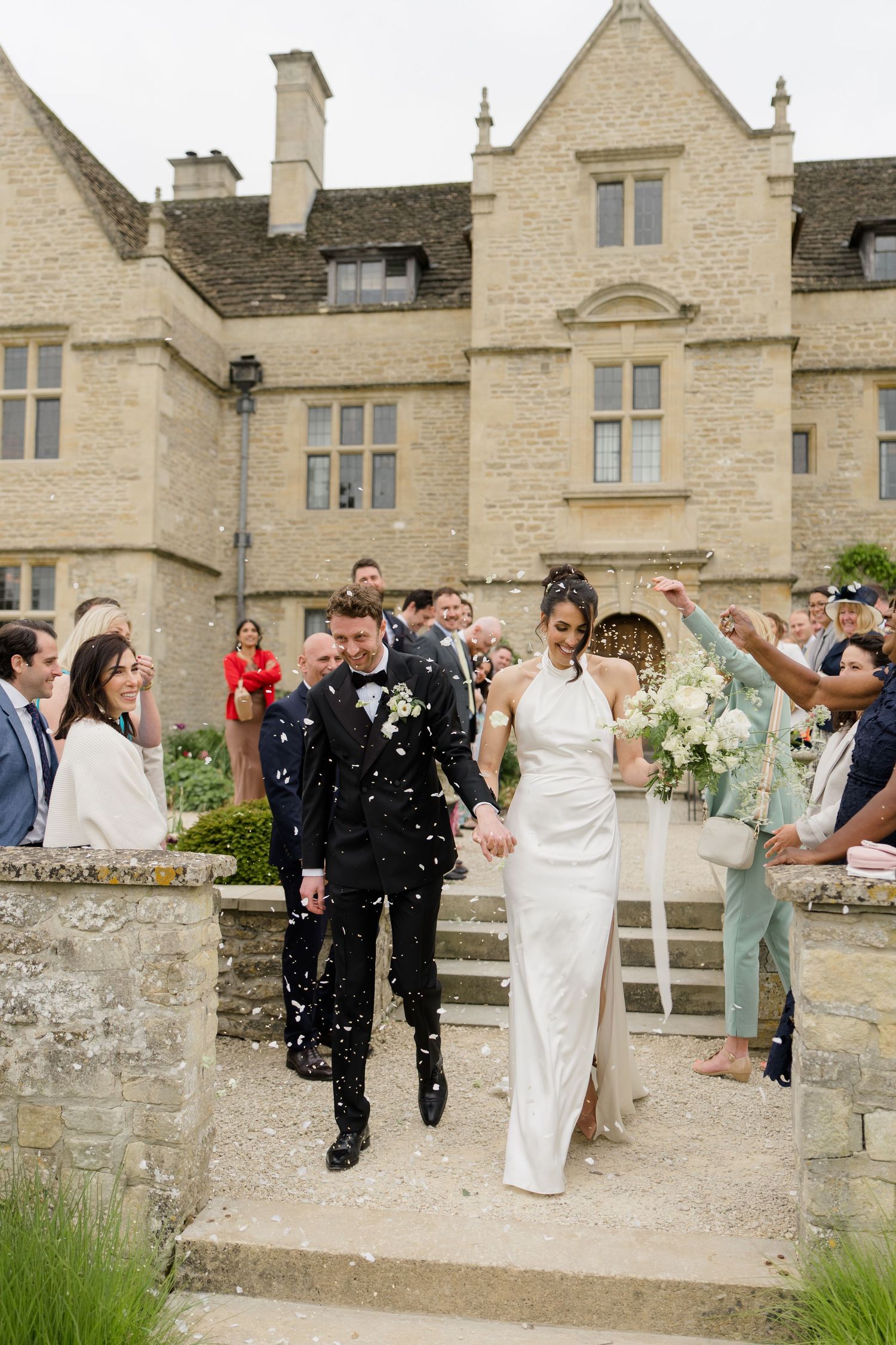 Smiling newlyweds exit through confetti outside a grand English manor house after wedding ceremony.