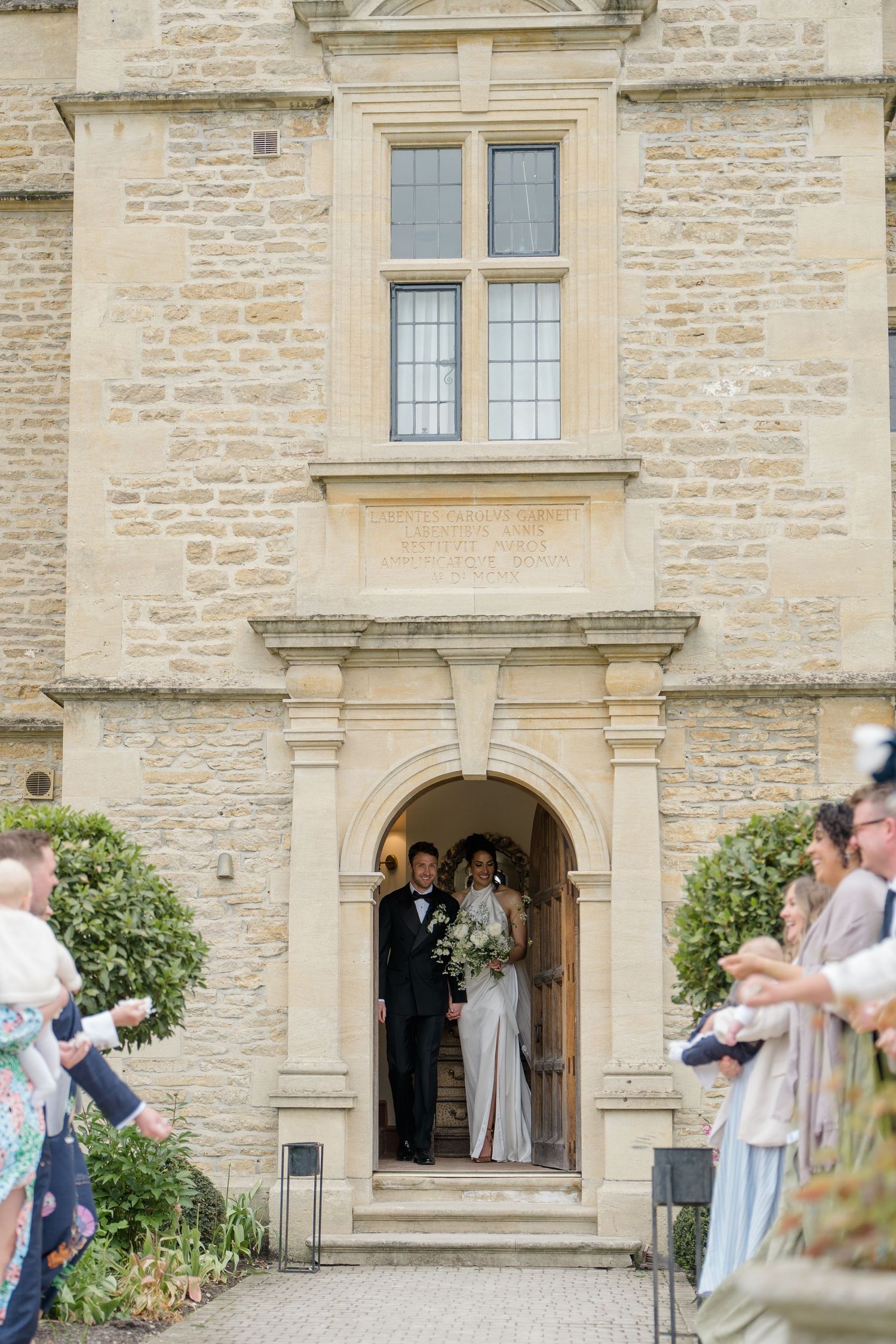 Bride and groom stepping out of an elegant stone manor entrance as guests line up to celebrate them.