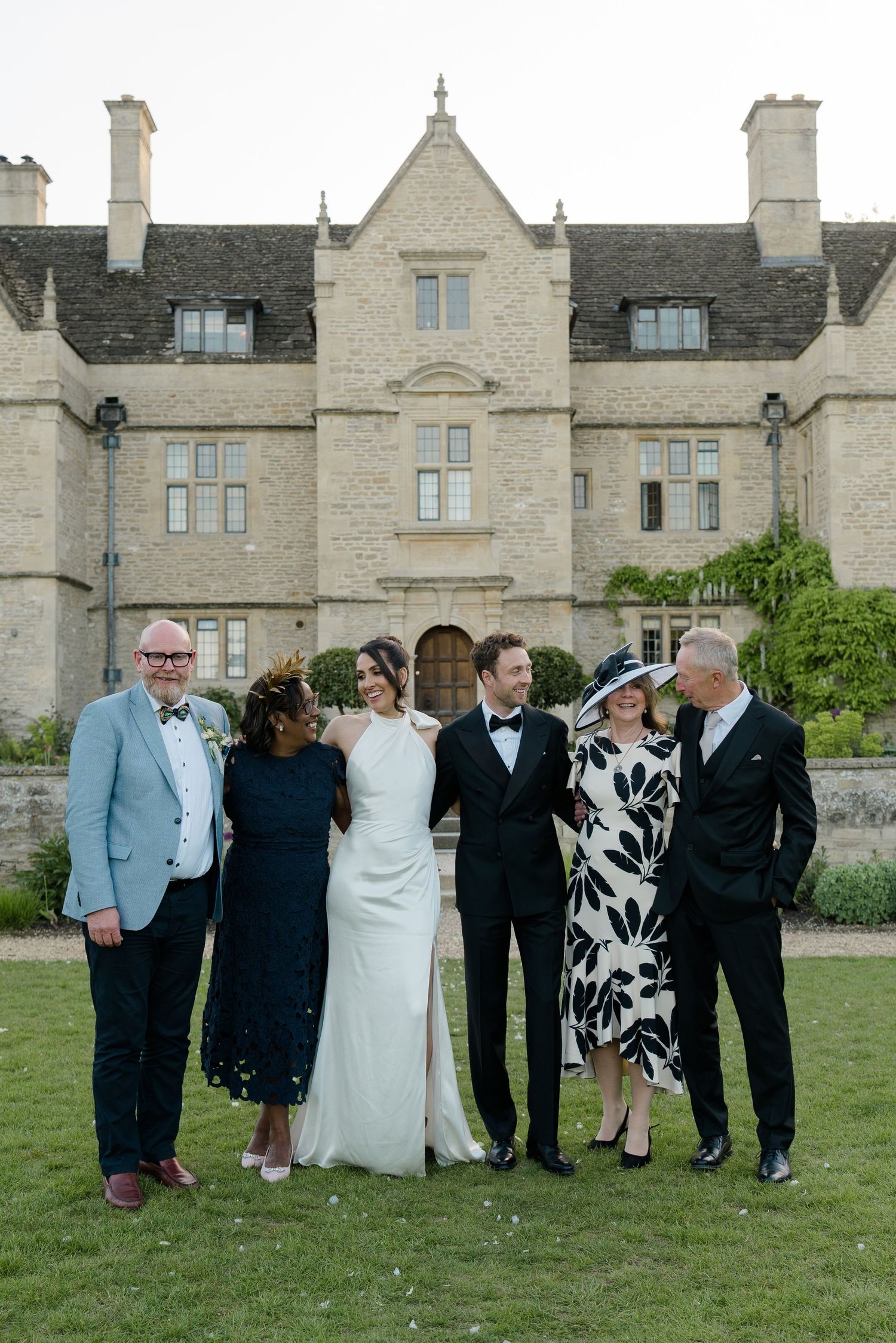 Wedding party of six posing in front of a grand stone manor house at dusk on the lawn.