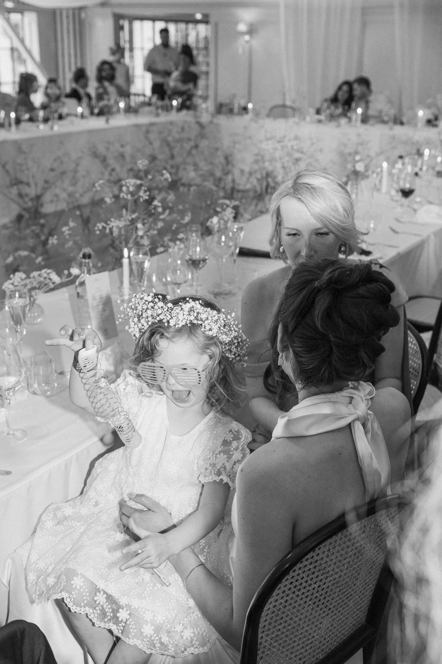 Black and white photo of flower girl in funky glasses entertaining bride at reception.