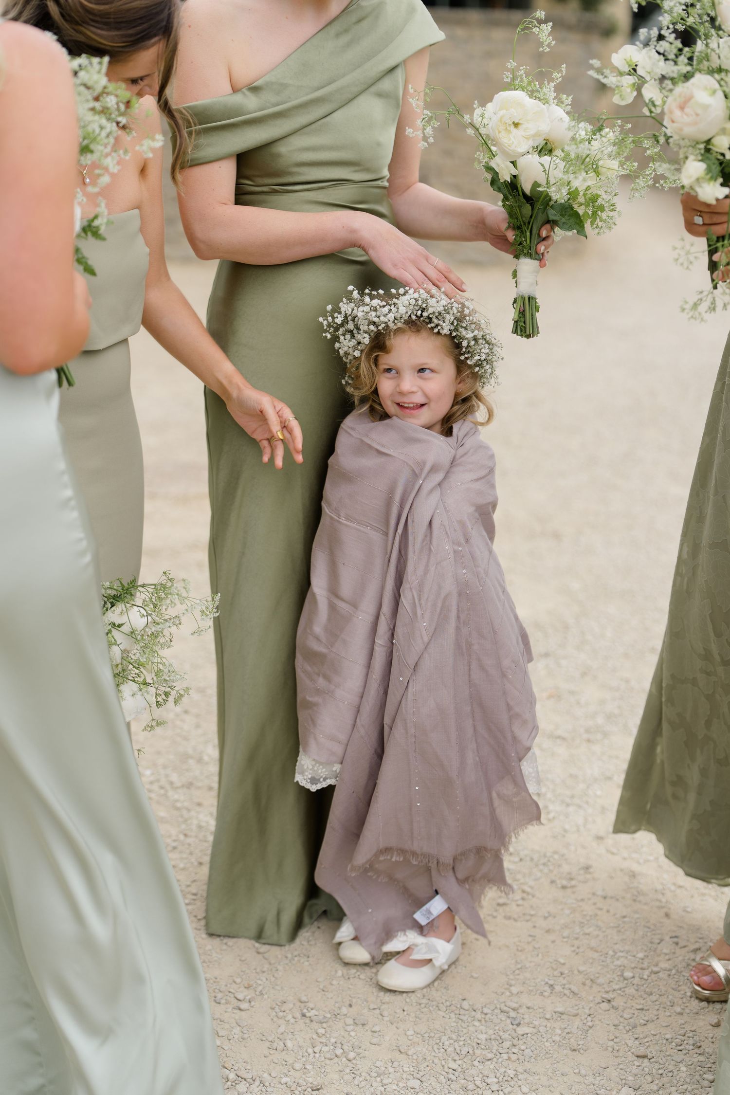 Adorable flower girl wearing floral crown wrapped in linen shawl standing with bridesmaids.