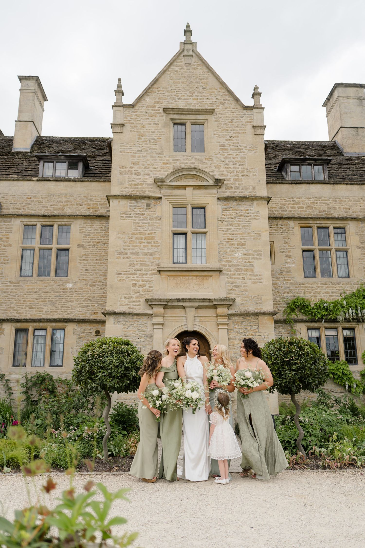 Bride in white halter gown with bridesmaids in sage green dresses outside a stone manor house.