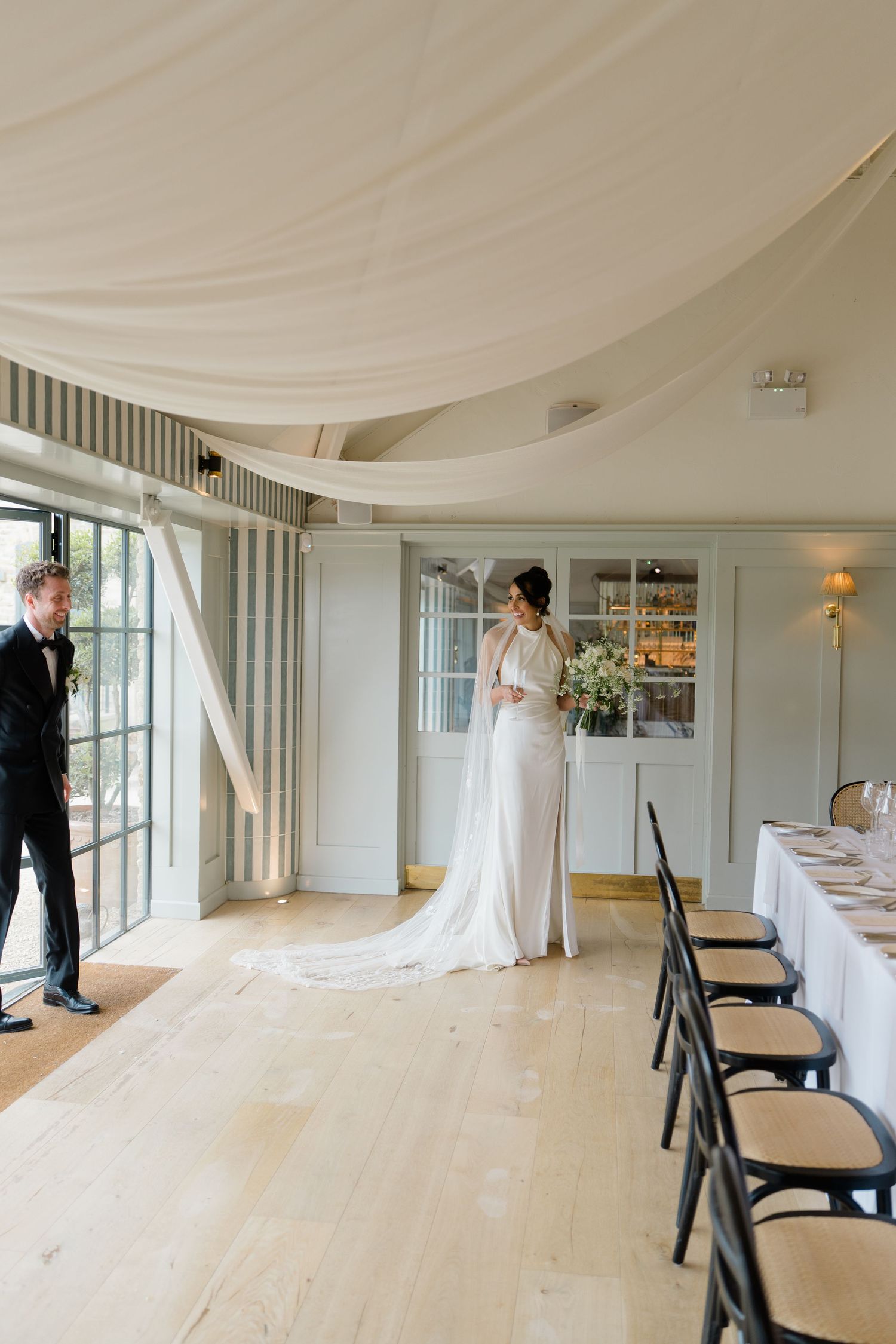 Bride in elegant white gown with train stands in beautifully draped reception hall as groom approaches.