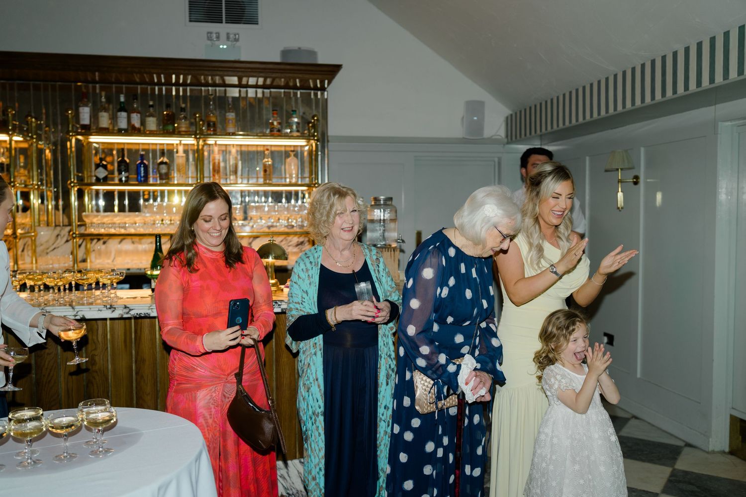 Wedding guests gather around elderly woman near bar as excited flower girl reacts with delight.