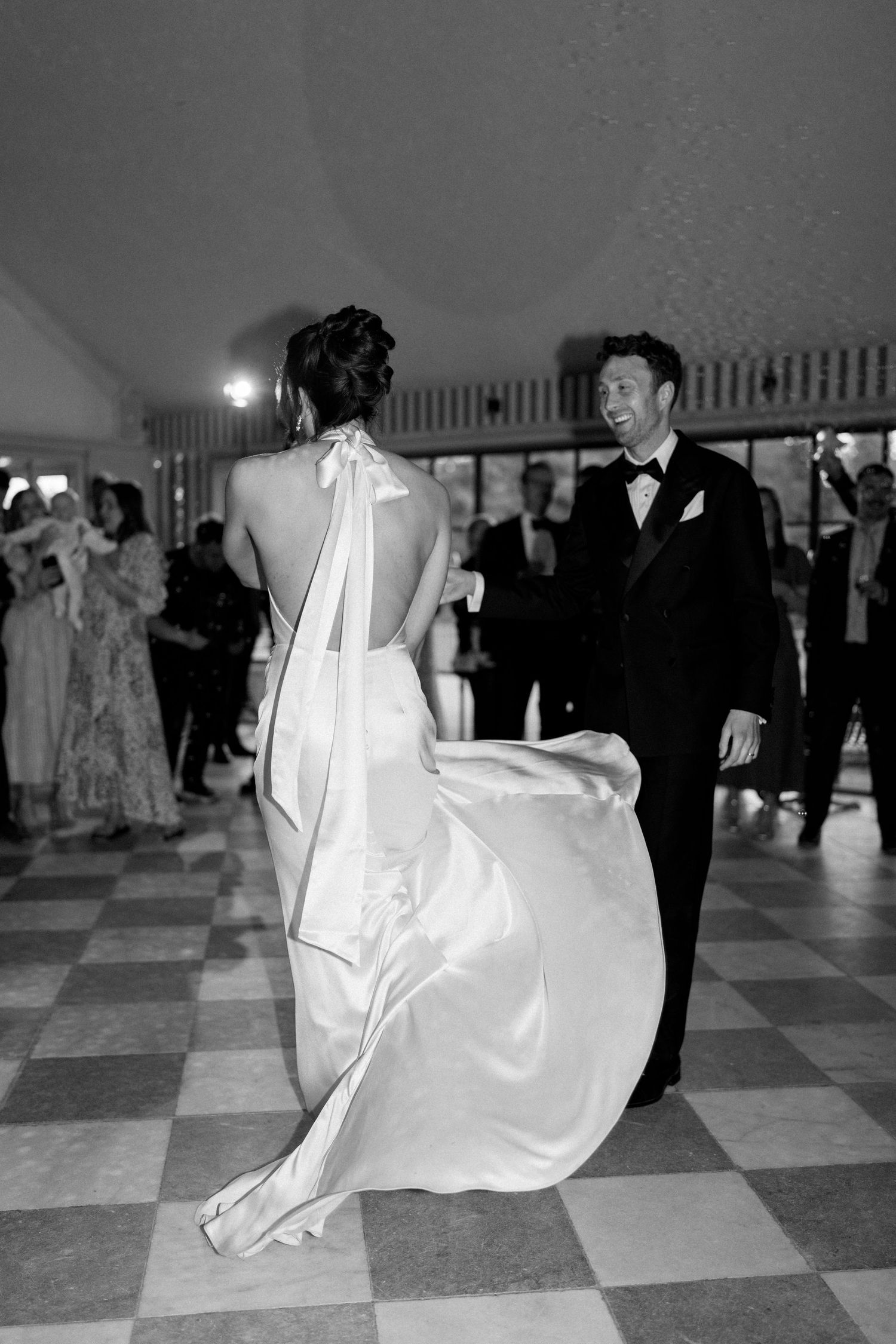 Bride in backless halter wedding gown twirls toward smiling groom on checkered ballroom dance floor.
