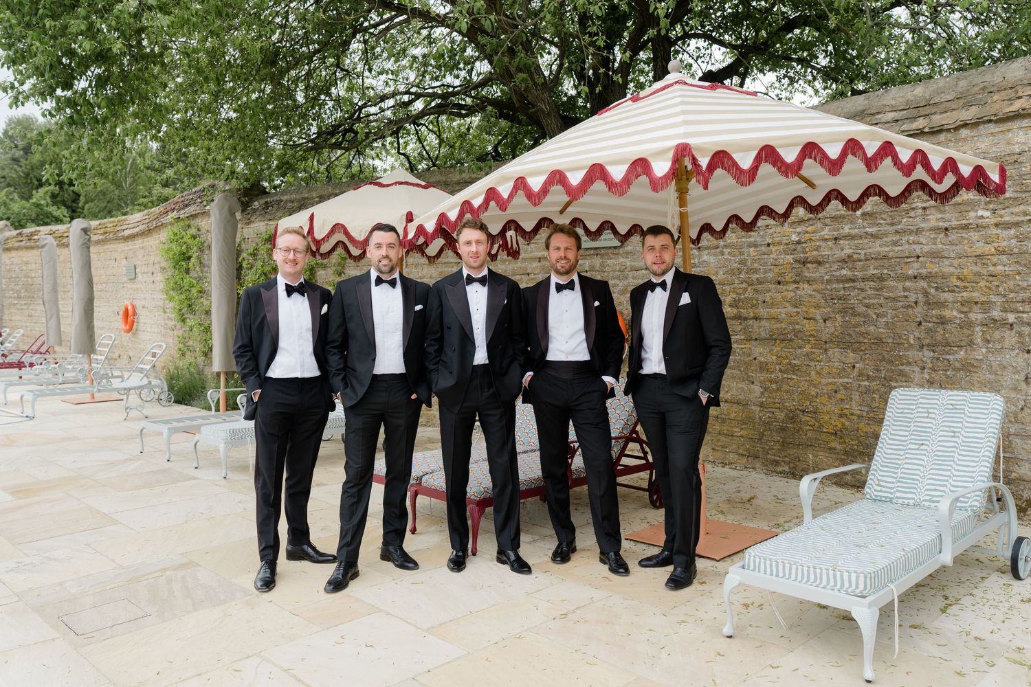 Five groomsmen in black tuxedos posing under a red and white striped umbrella on a stone patio.