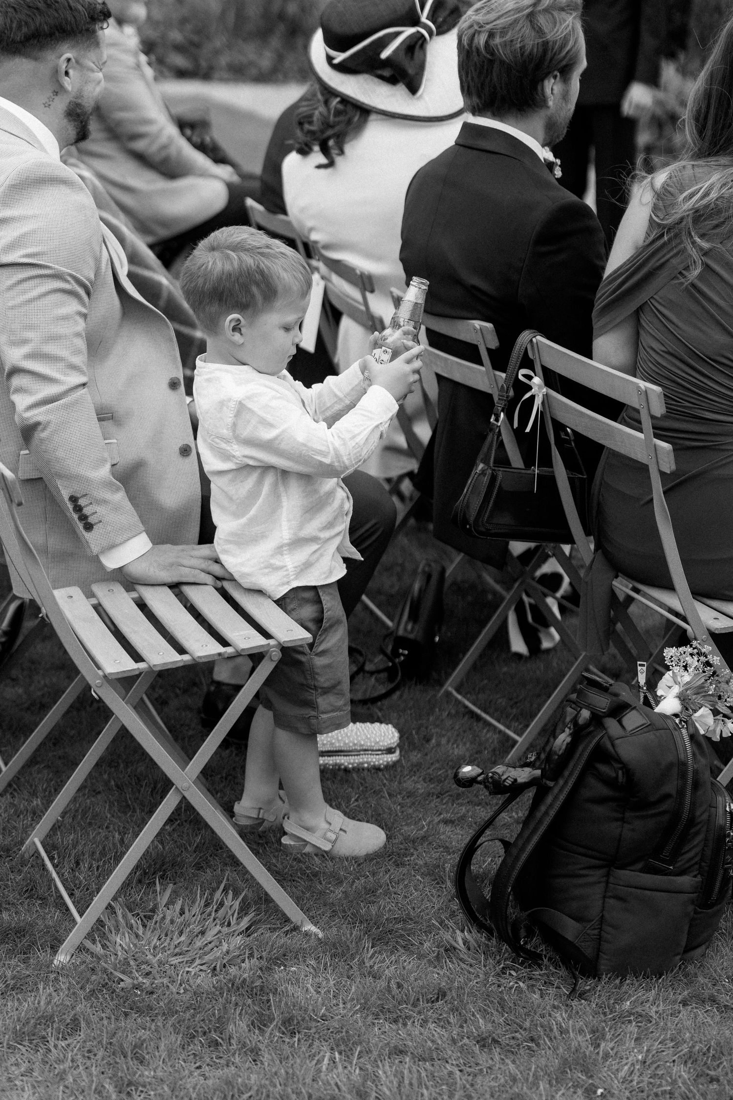 Young toddler boy standing on chair holding bottle at outdoor garden wedding ceremony.
