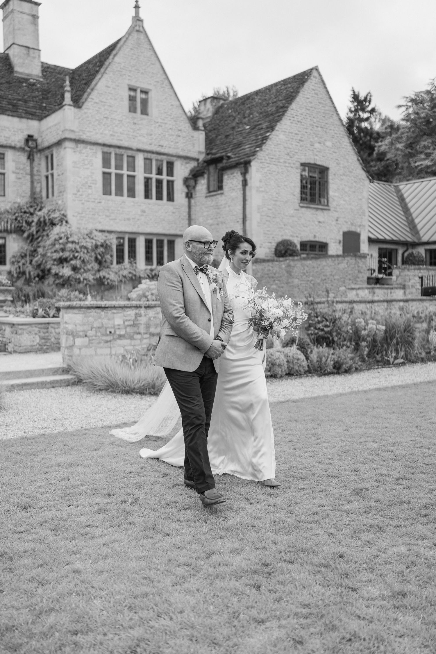 Bride and escort walk arm in arm across lawn in front of a classic English country manor house.