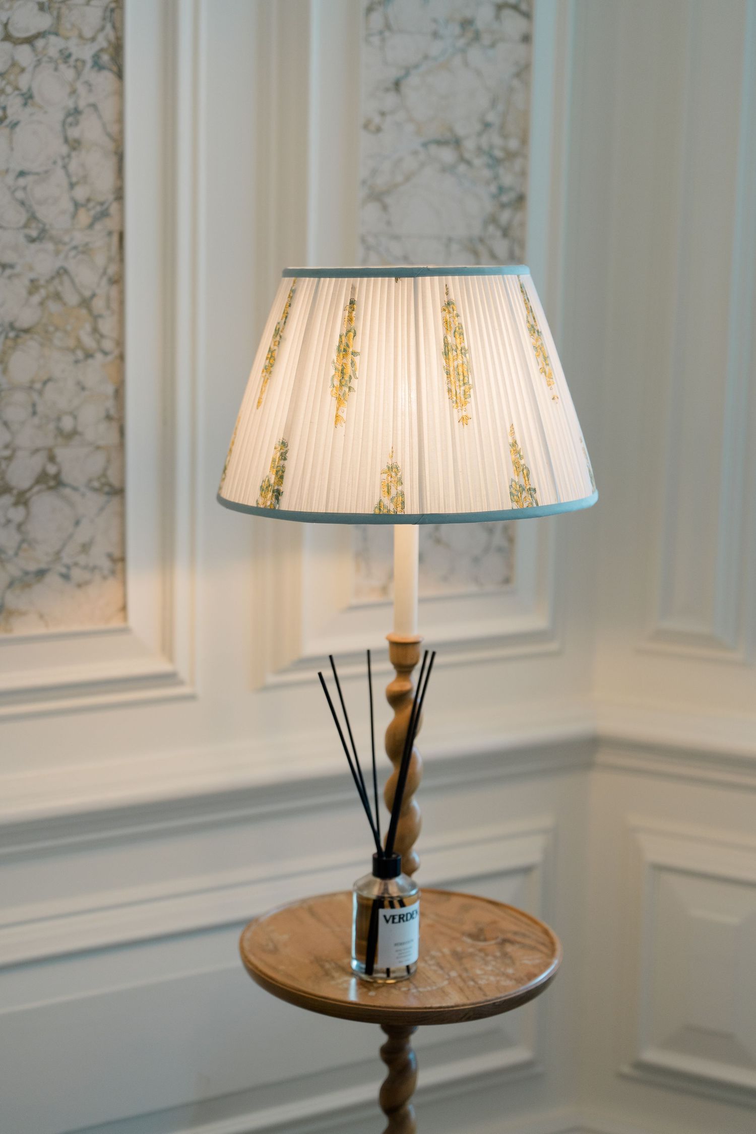 Elegant pleated lamp shade with blue trim on wooden side table with reed diffuser against marble wallpaper.