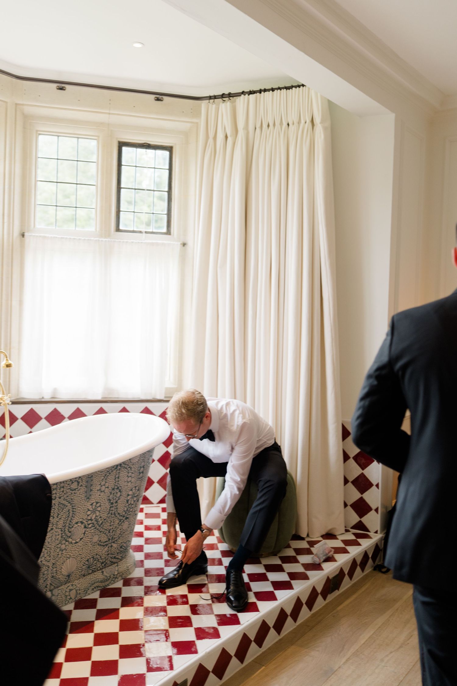 Groom putting on shoes in elegant bathroom with red checkered tile floor before wedding ceremony.
