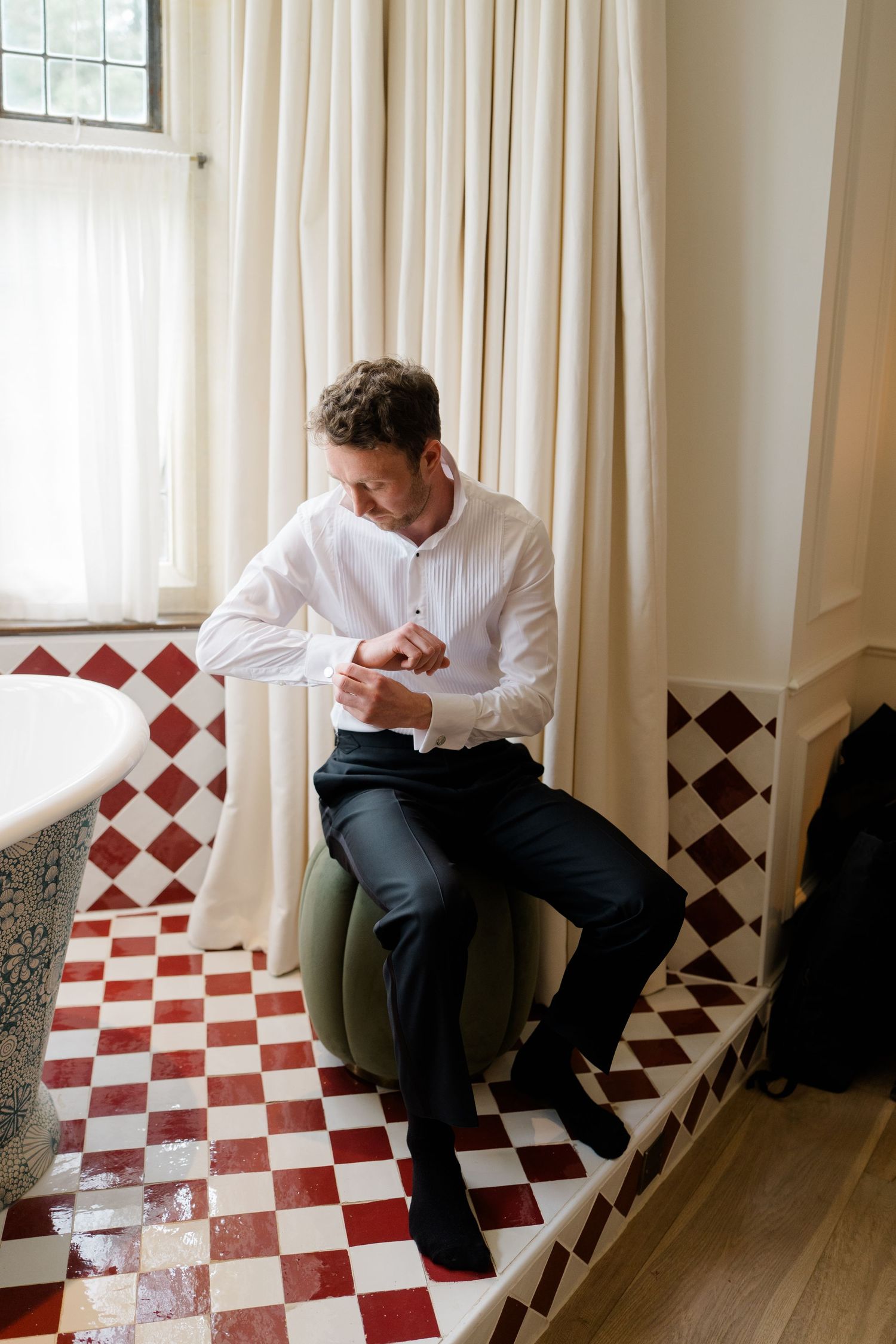 Groom in white shirt fastening cufflinks seated on ottoman in checkered tile bathroom.