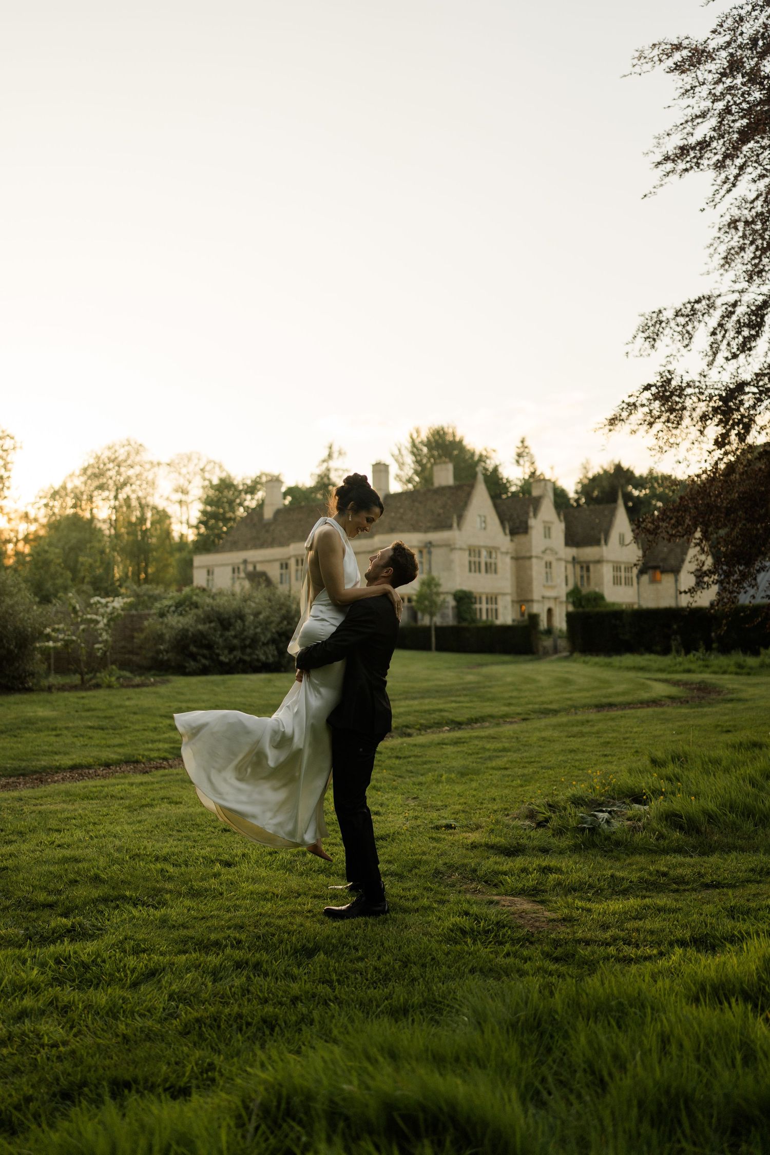 Groom lifts bride in flowing white dress at golden hour in lush green gardens of a historic manor house.