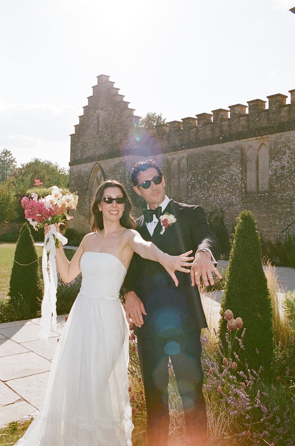 Bride and groom in sunglasses pose joyfully outside a stone castle venue, bride holding pink bouquet on sunny wedding day.