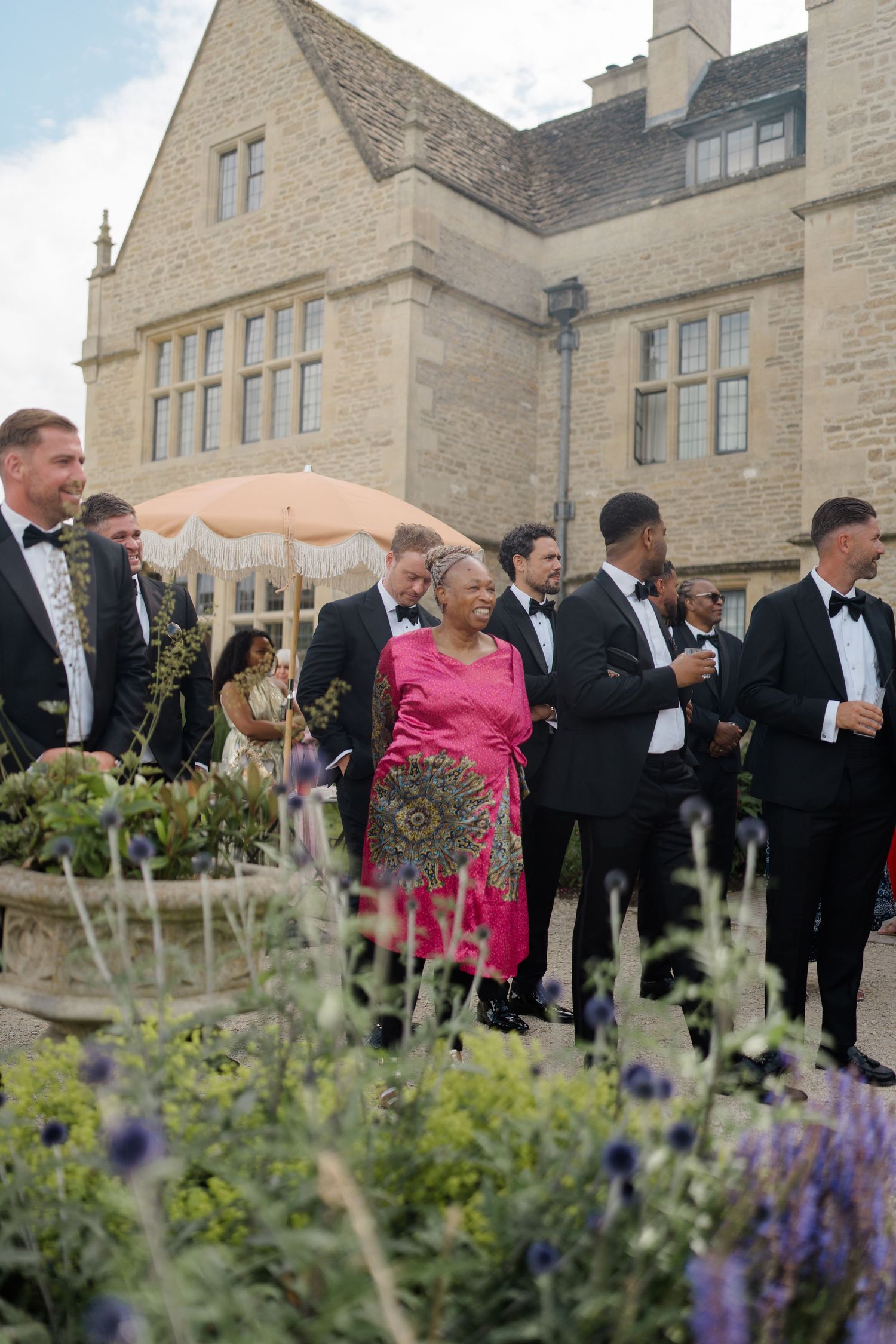 Wedding guests in formal attire mingling outside historic stone manor house with garden flowers in foreground.