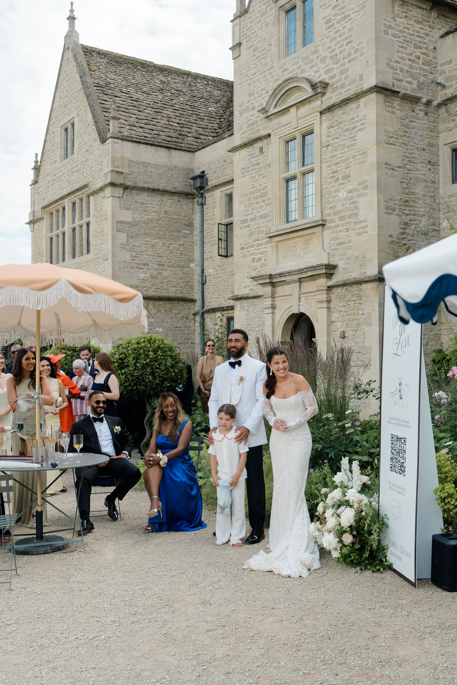 A bride and groom stand with a young page boy outside a grand stone manor house during their wedding ceremony.