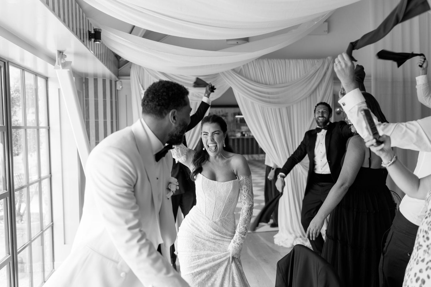 Black and white photo of joyful bride and groom making grand entrance to wedding reception with cheering guests.