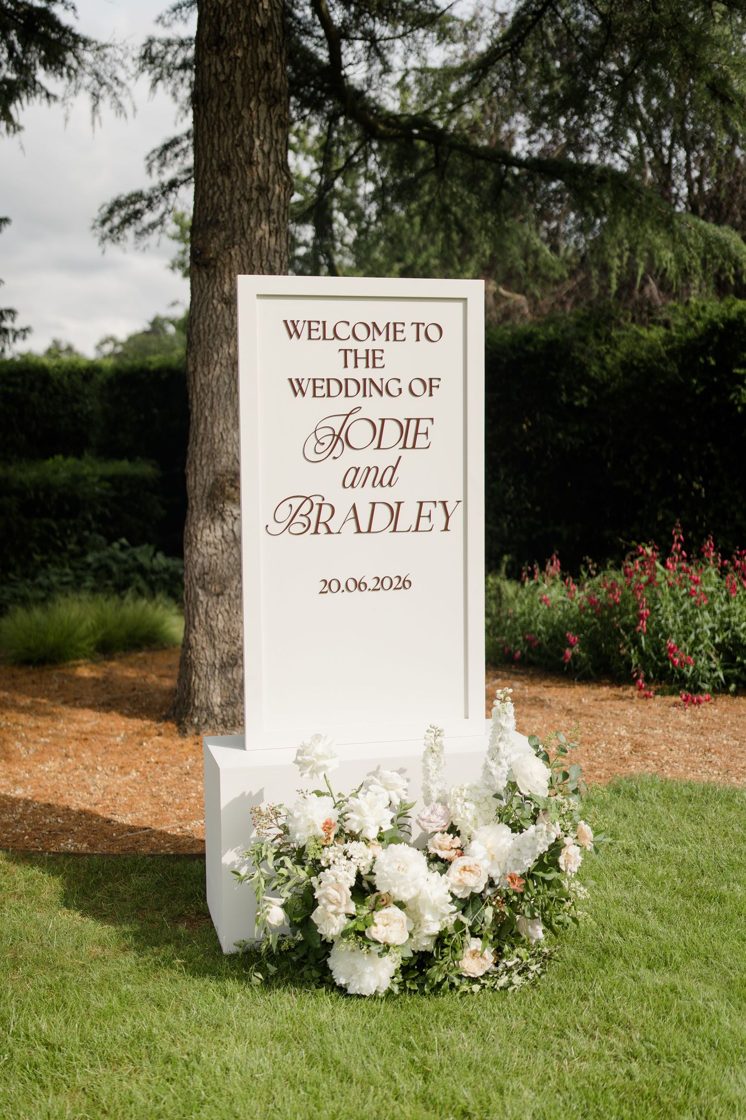 Elegant white wedding welcome sign for Jodie and Bradley dated 20.06.2026 adorned with white floral arrangements.