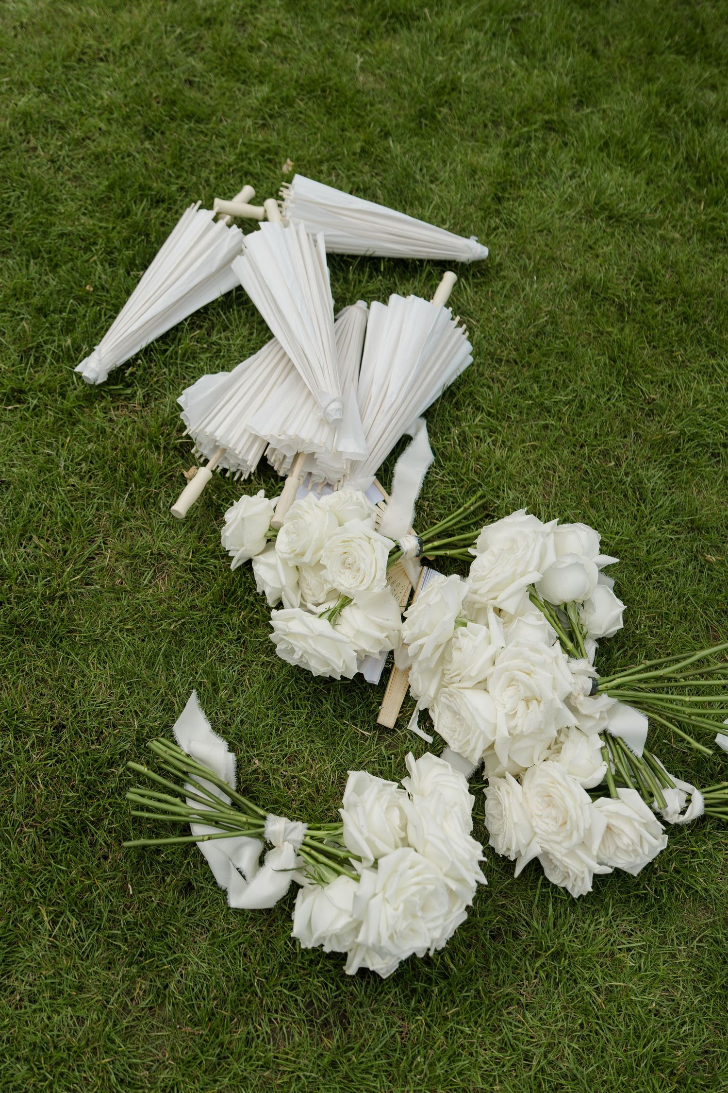 White wedding bouquets of roses and folded parasols arranged on green grass before ceremony.