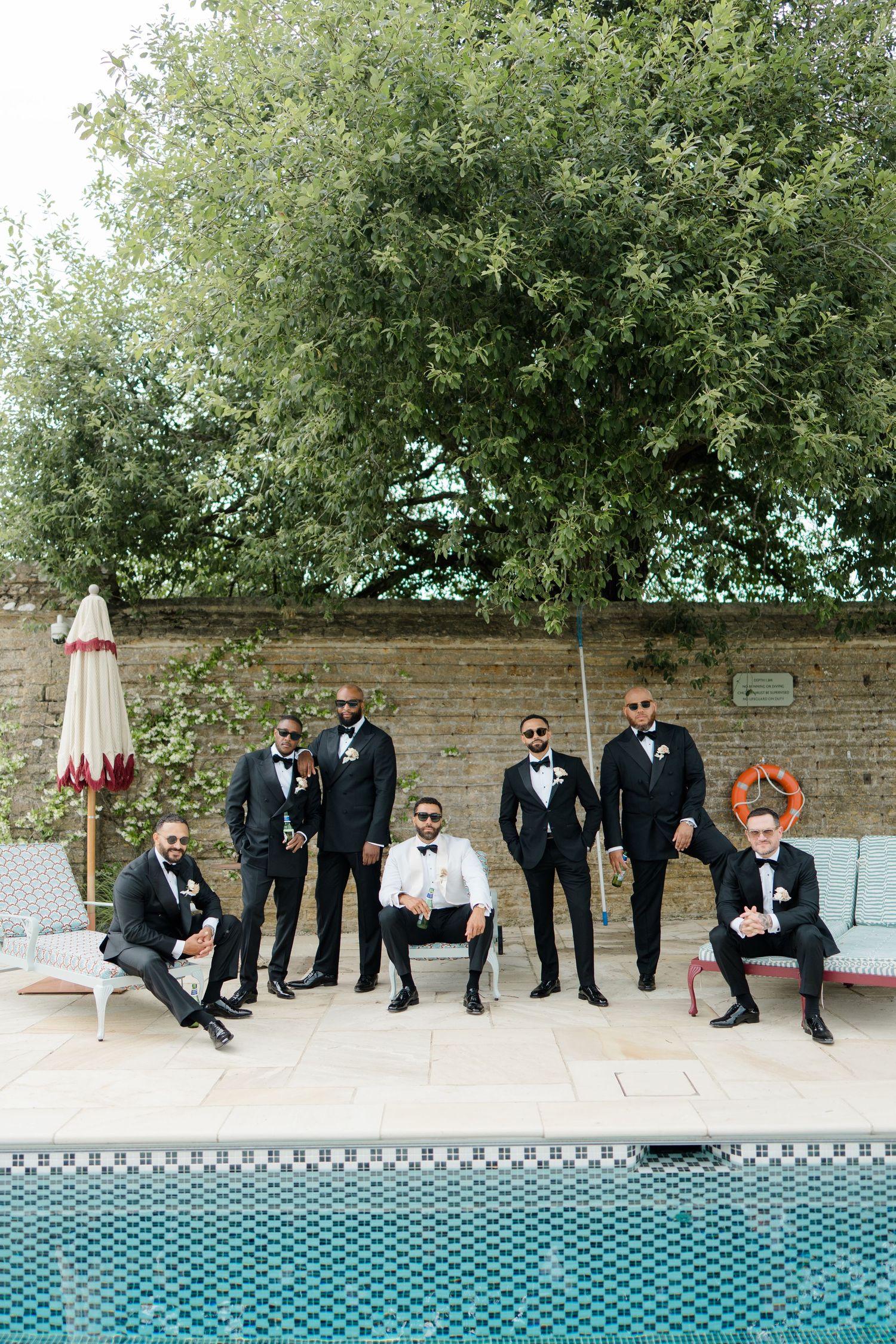 Groom and groomsmen in black tuxedos posing by outdoor swimming pool at wedding venue.