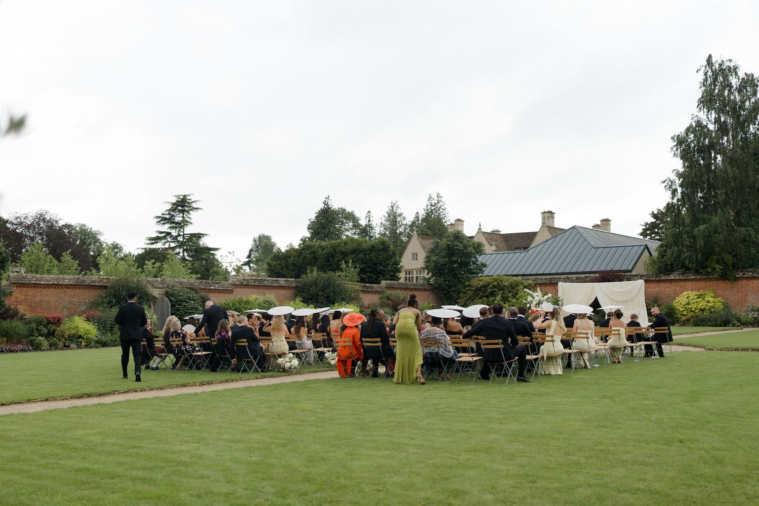 Guests seated on white chairs at elegant outdoor wedding ceremony in lush green garden setting.