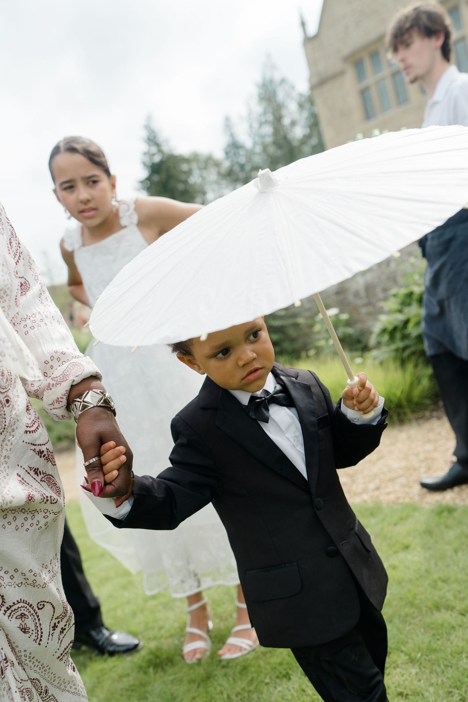 Adorable young ring bearer in black tuxedo holds a white parasol umbrella at an outdoor wedding ceremony.