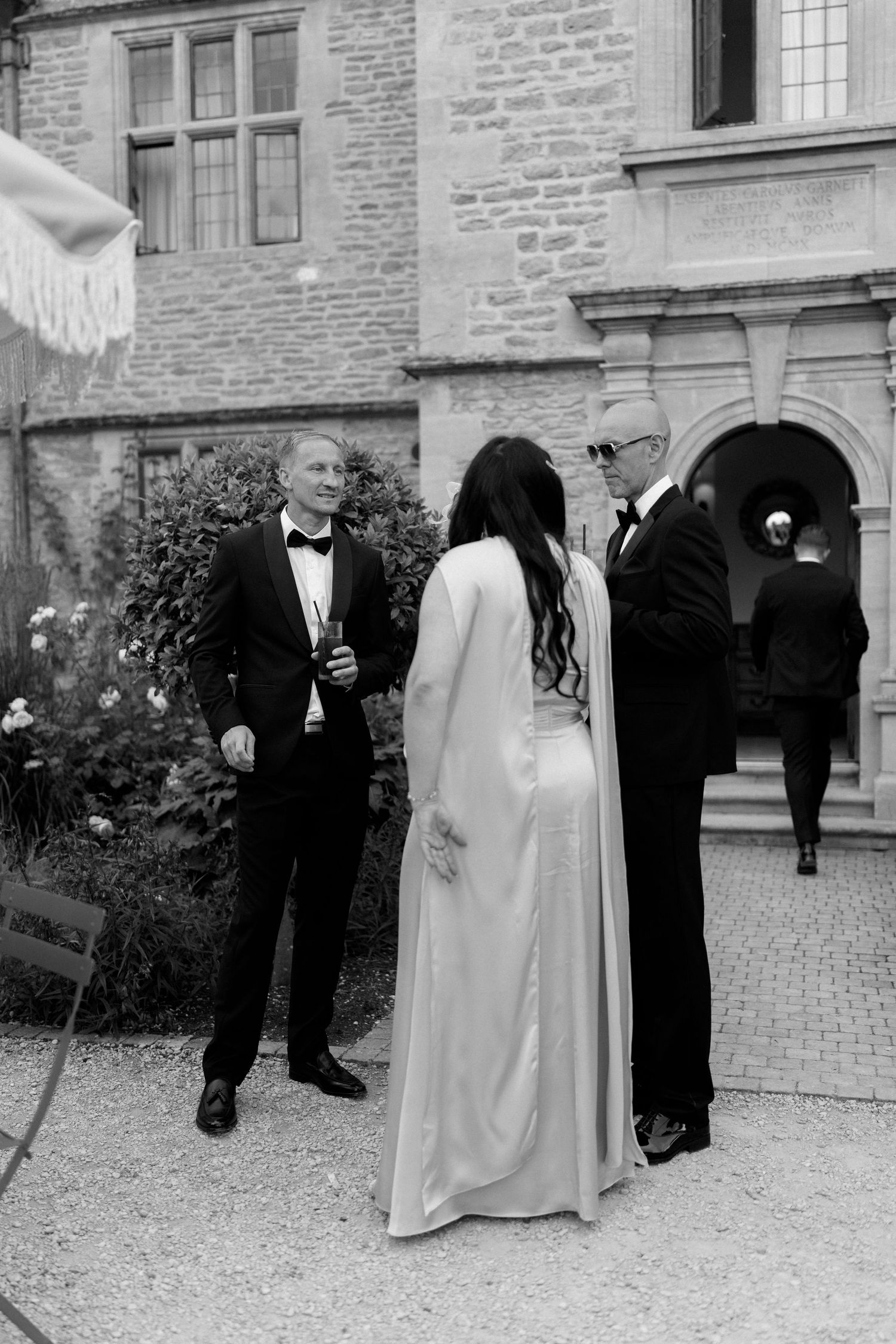 Groom in tuxedo chats with bride in white cape gown and guest outside stone venue in black and white.