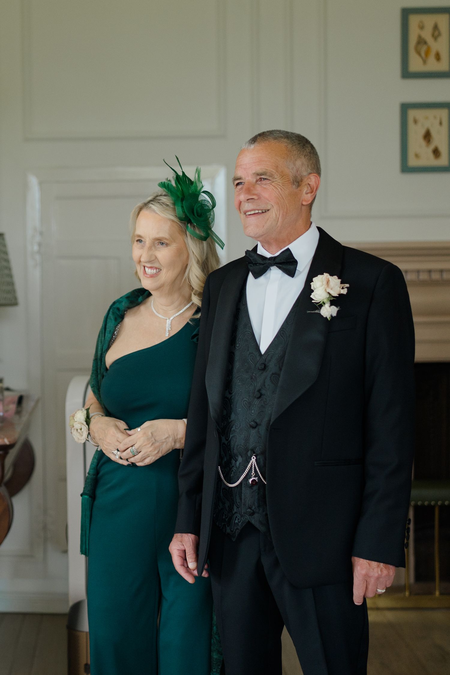 Elegant couple in formal wedding attire, woman in emerald green jumpsuit with feathered fascinator.